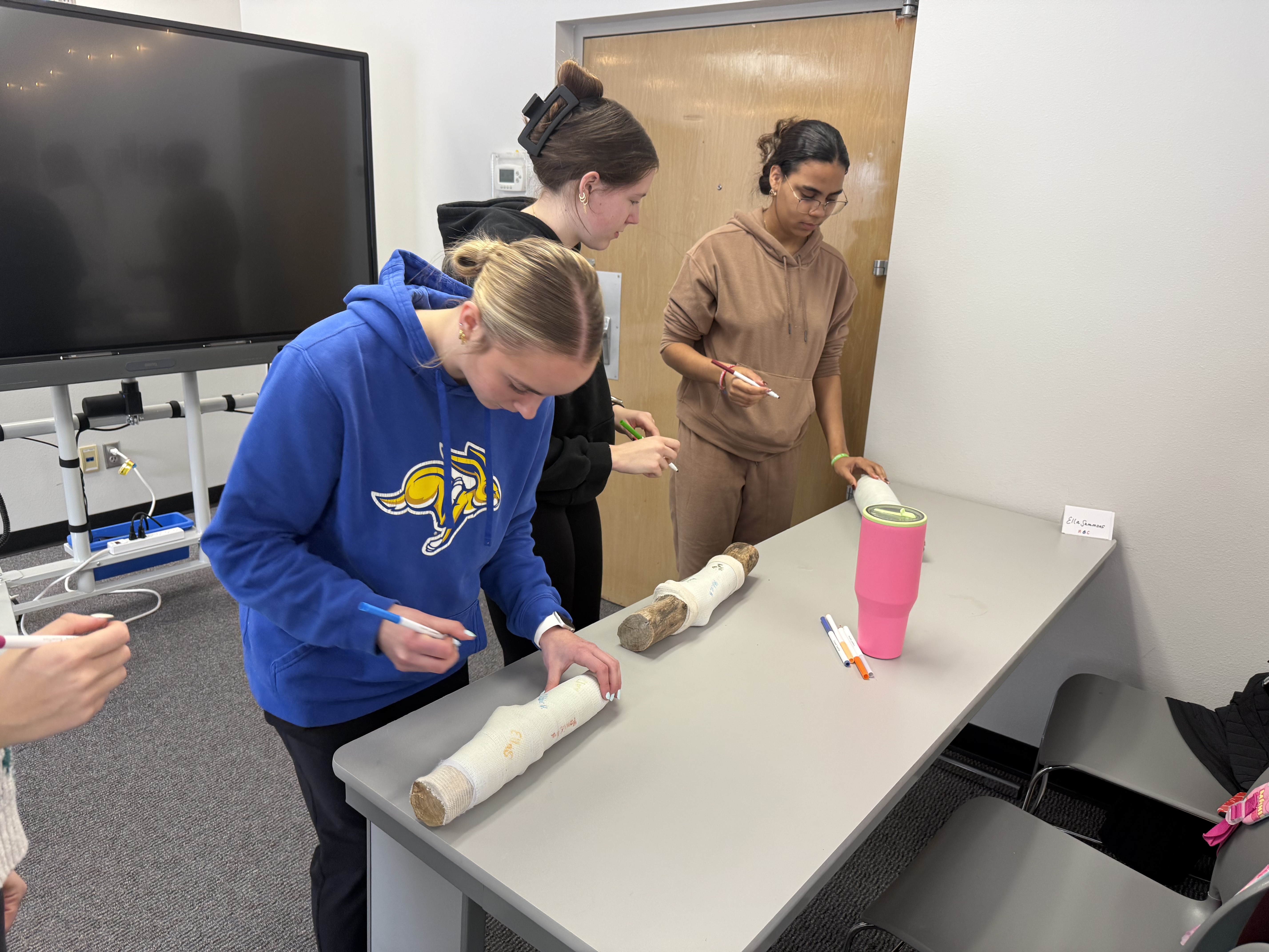 The image shows several people in a medical or healthcare setting, with one person wearing a blue sweatshirt and another wearing a beige outfit, all engaged in some kind of activity or task.