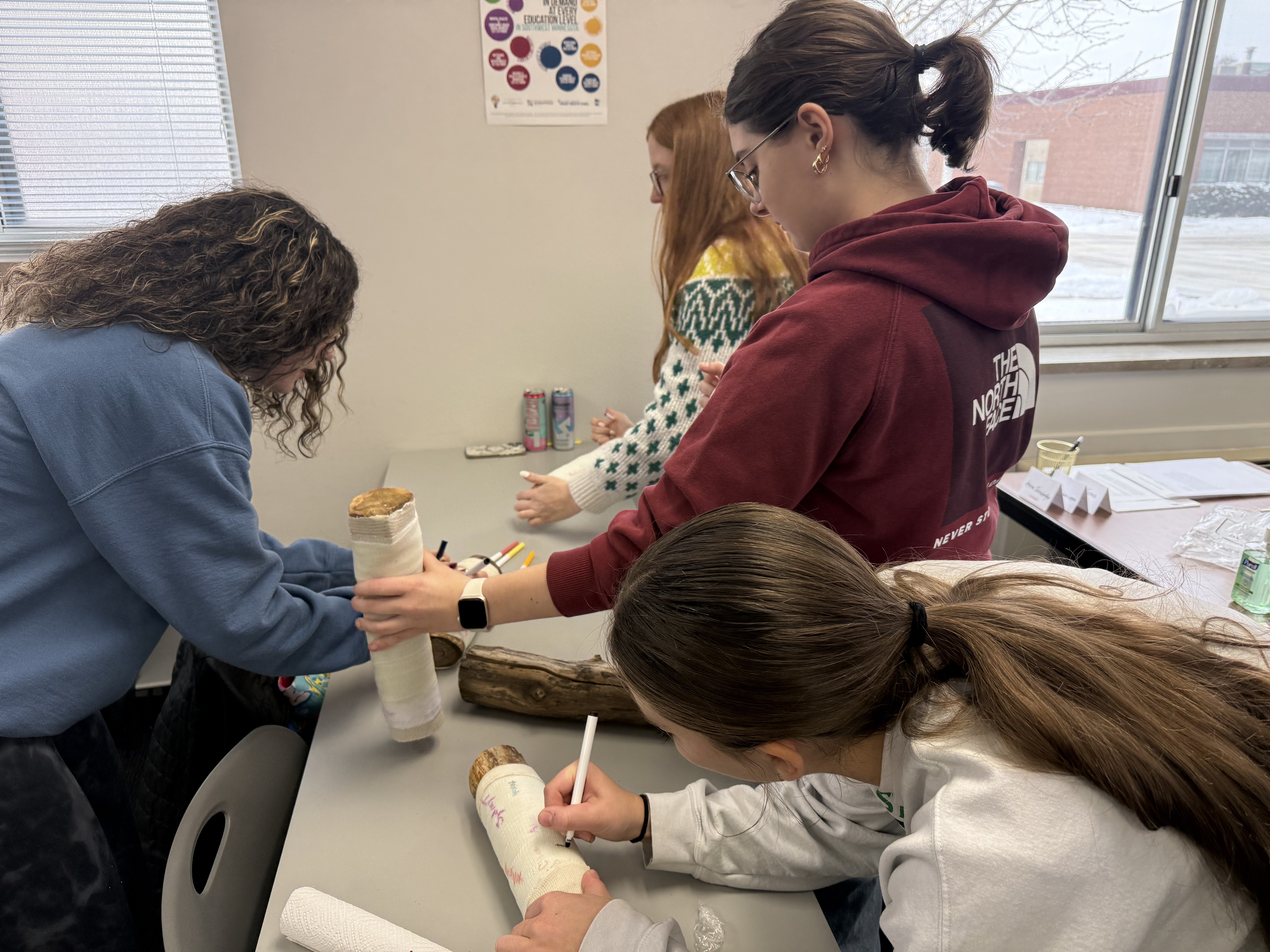 A group of young people, likely students, are gathered around a table, engaged in some kind of collaborative activity or project. The background suggests a classroom or study environment, with various decorations and materials visible.