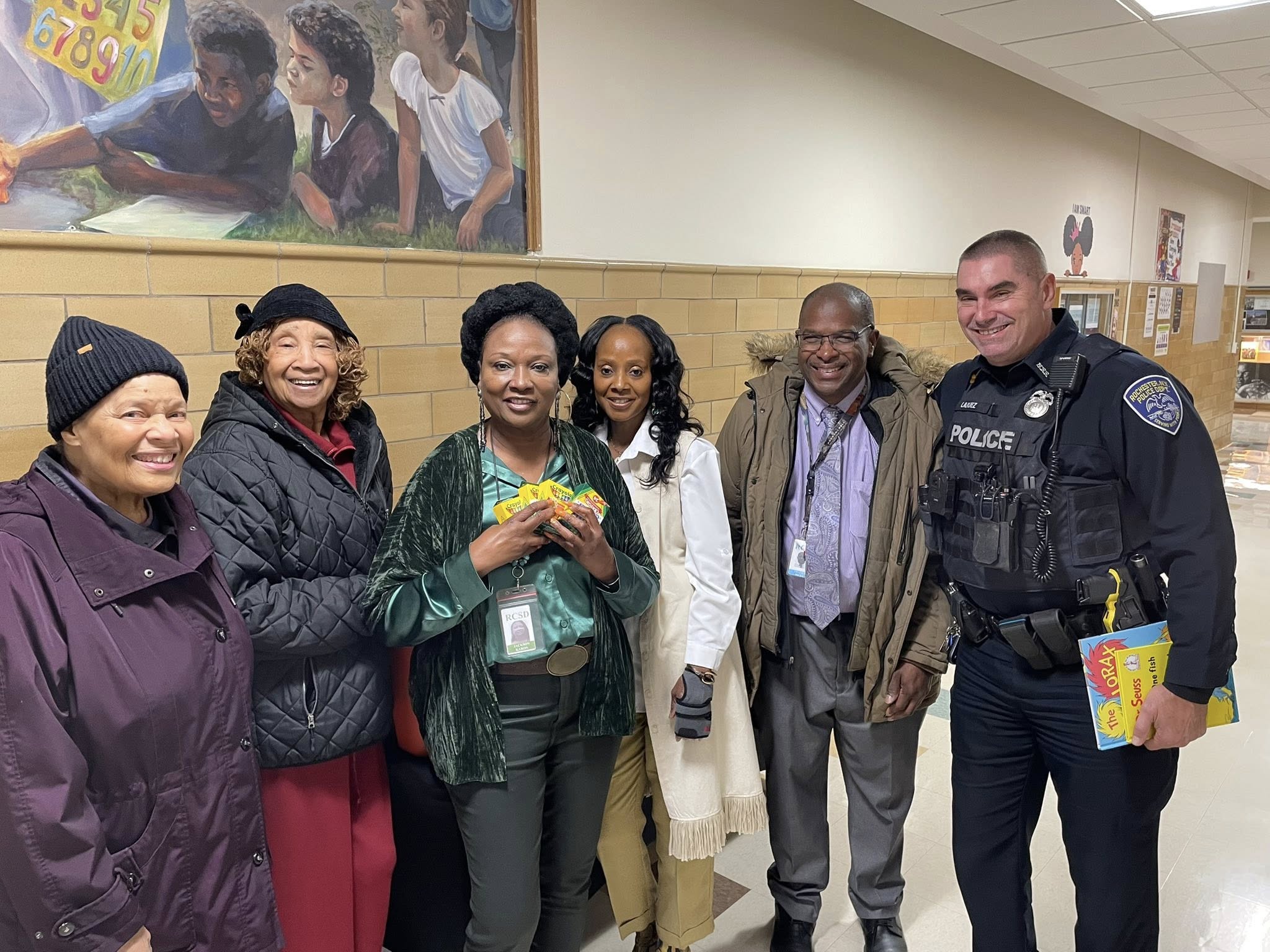 The image shows a group of people, including a police officer, standing together in what appears to be a school or community setting, with a large mural or artwork visible in the background.