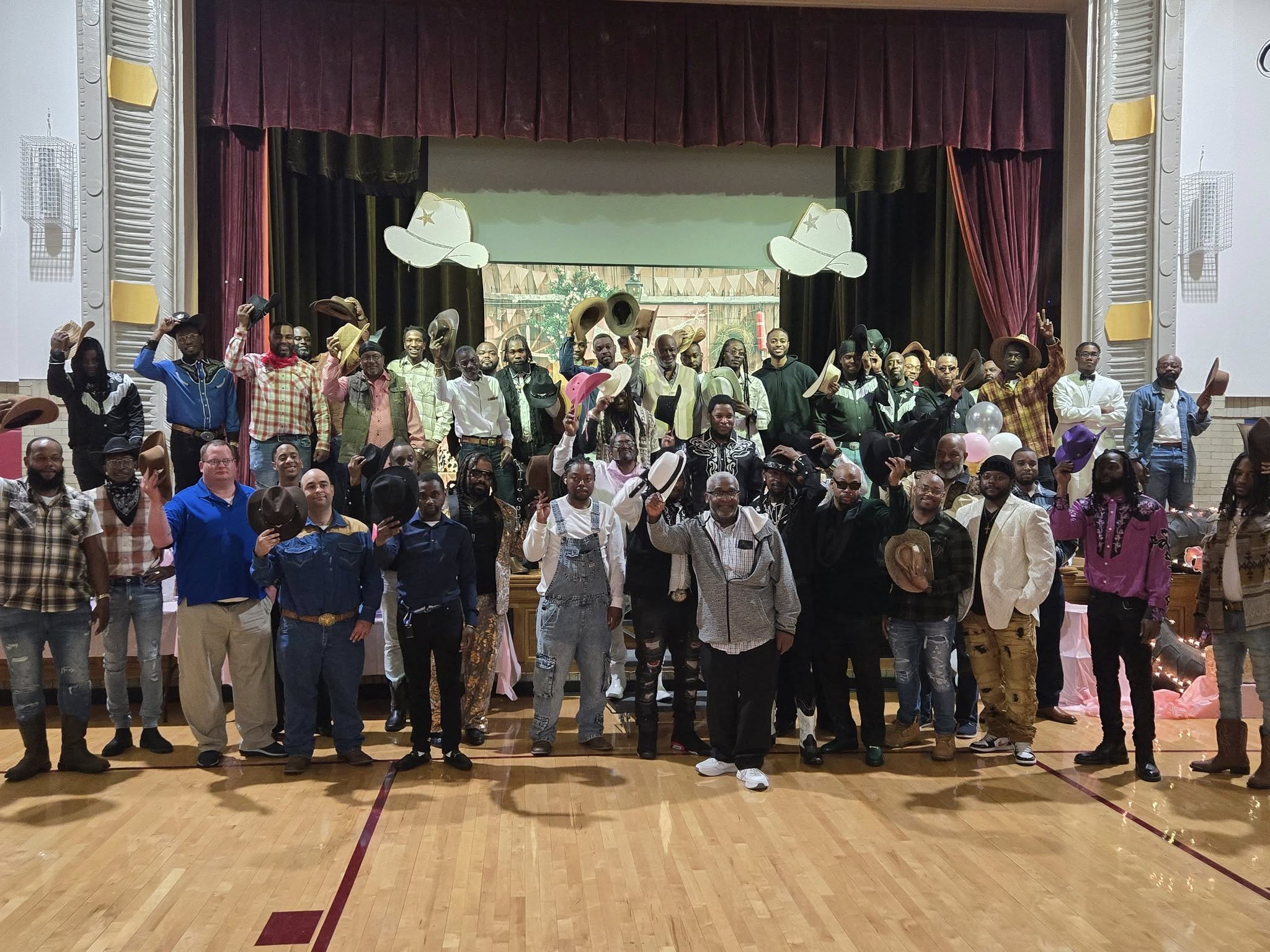 A large group of people, both young and old, gathered together in what appears to be a stage or auditorium setting, with a curtain backdrop and decorative elements visible.