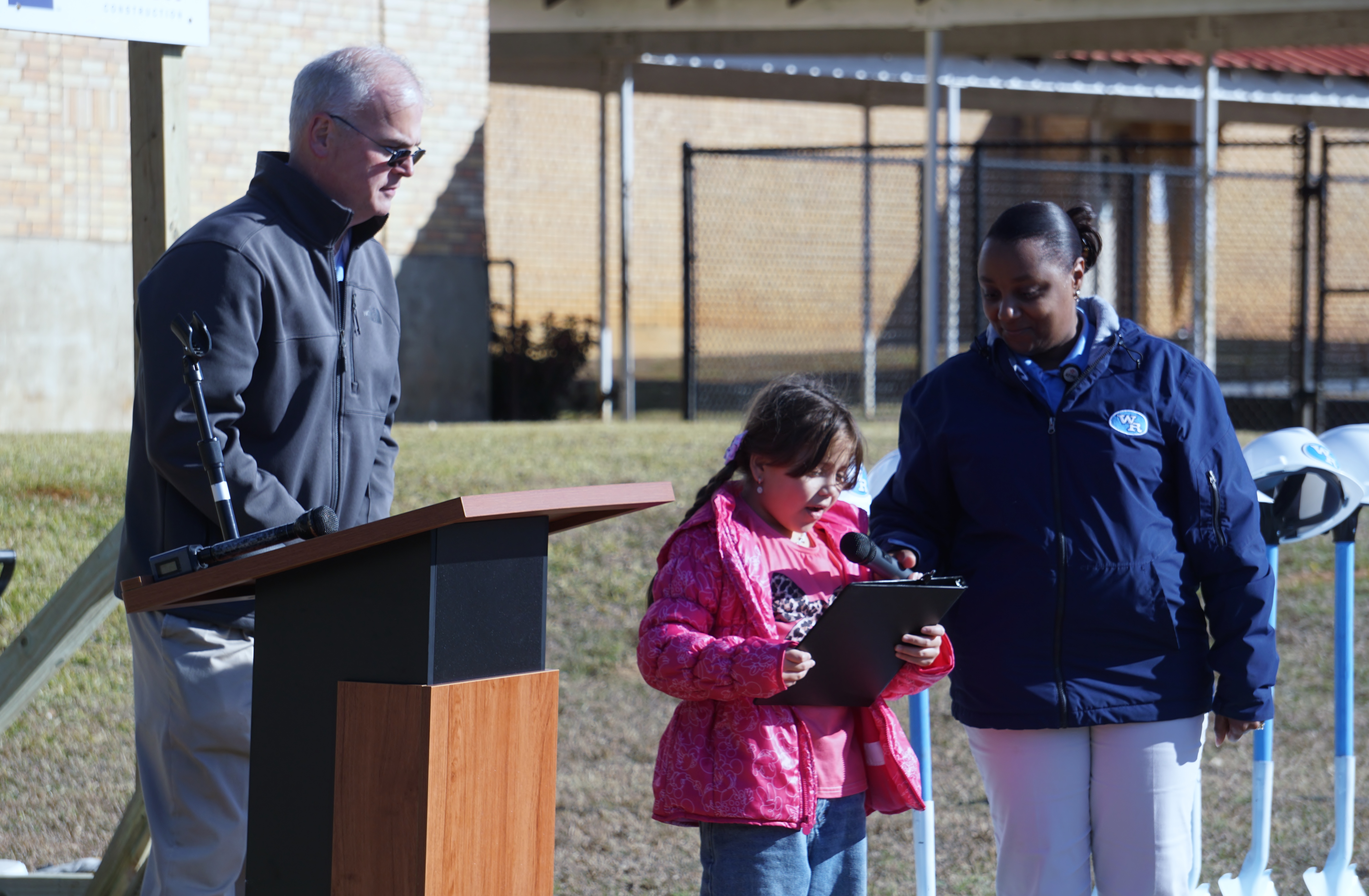 The image shows a man standing at a podium, with a woman and a young girl standing next to him, all of them dressed in casual attire.
