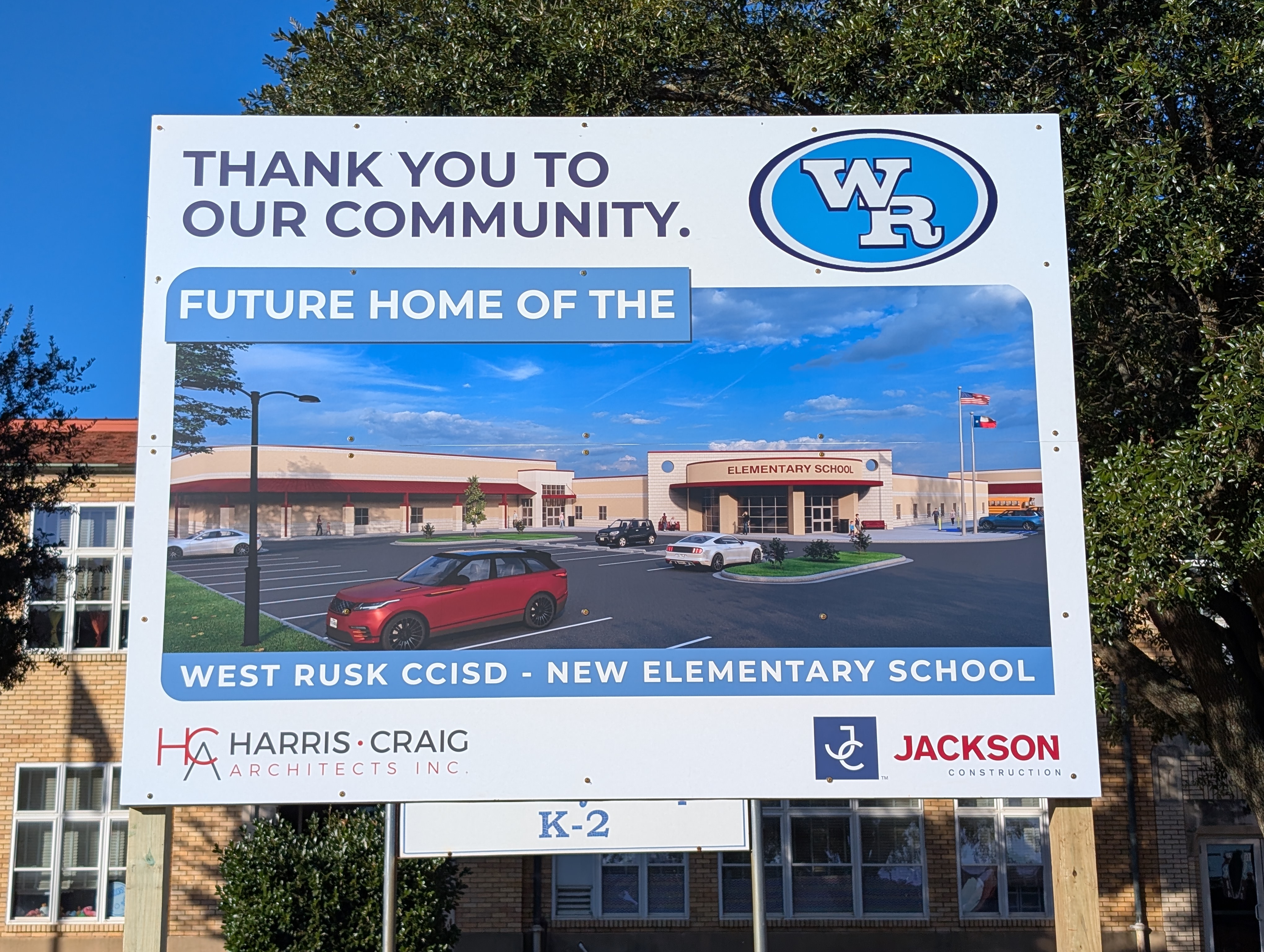 The image shows a sign thanking the community for their support, with a rendering of a new elementary school building in the background.