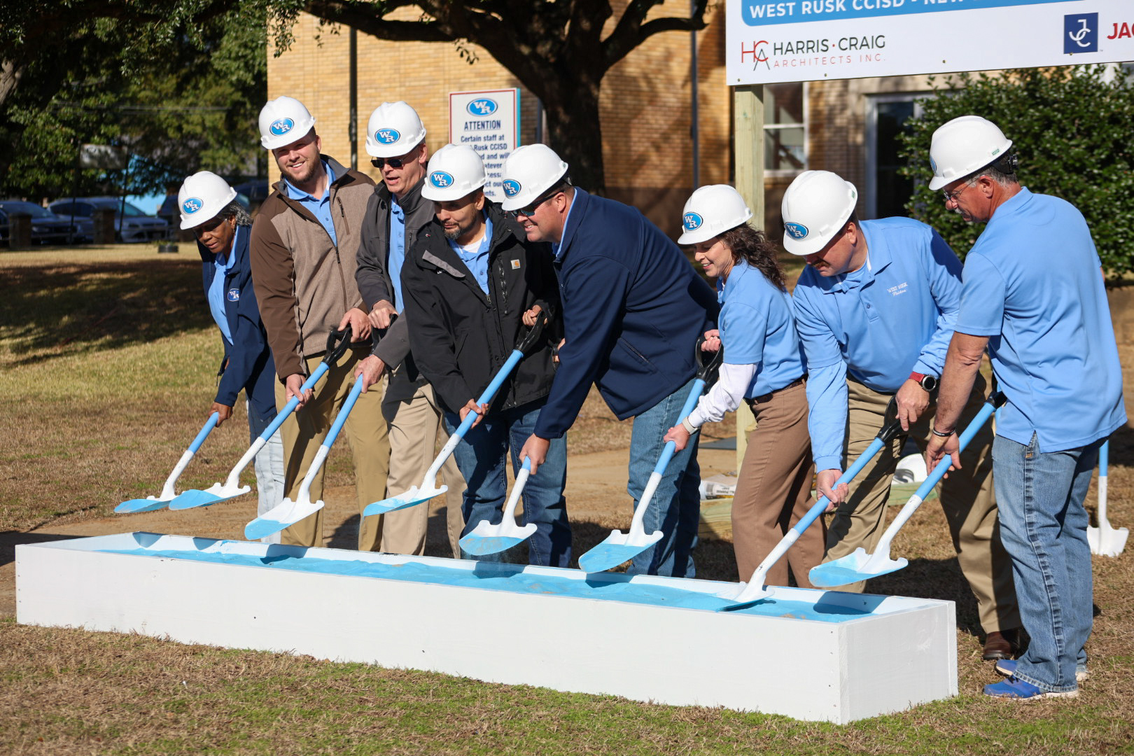 A group of people in hard hats and construction attire are gathered around a ceremonial groundbreaking event, with a construction site and signage visible in the background.