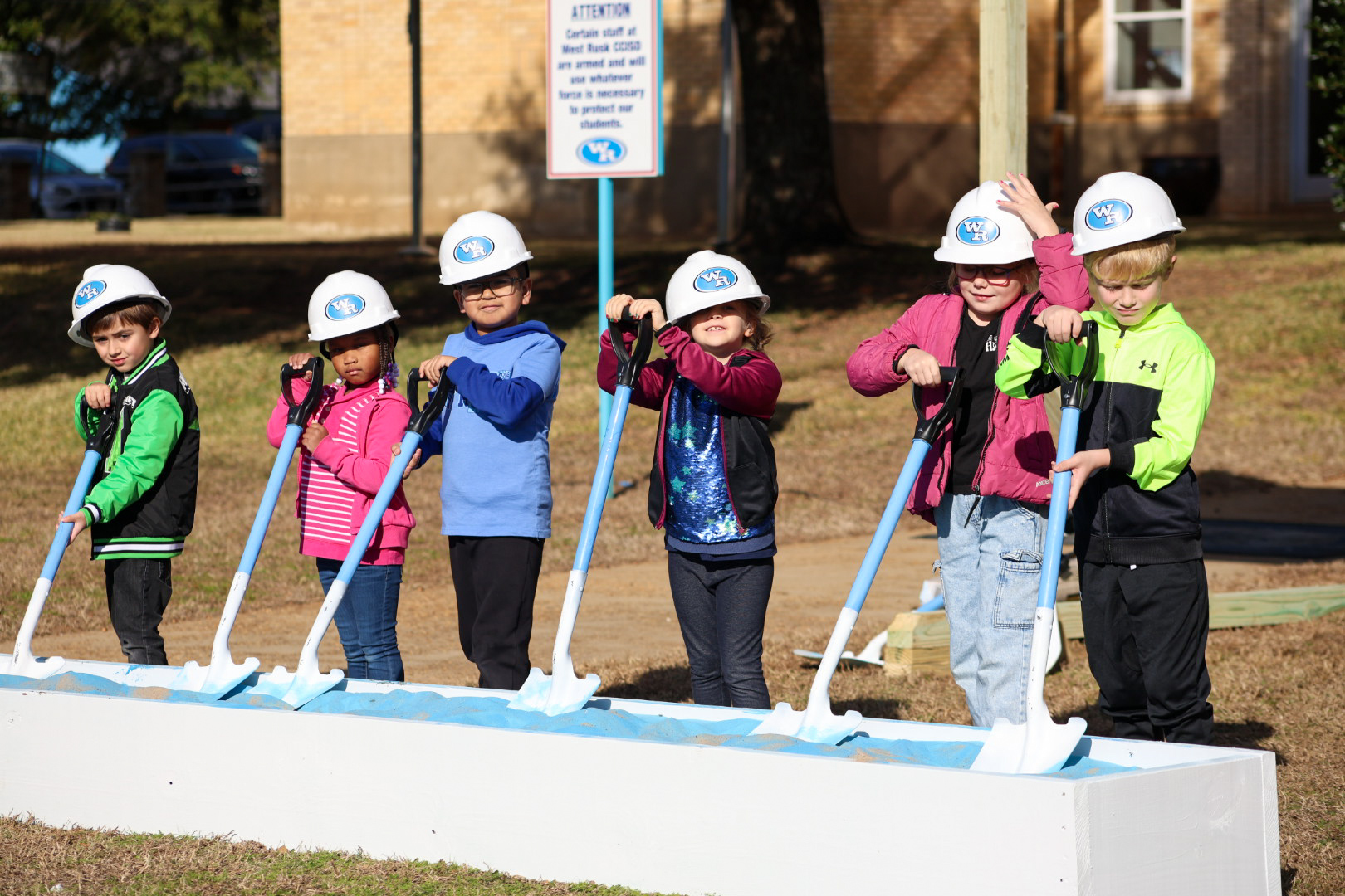 A group of children wearing hard hats and safety vests stand in front of a building, suggesting they are participating in a construction or engineering-related activity.