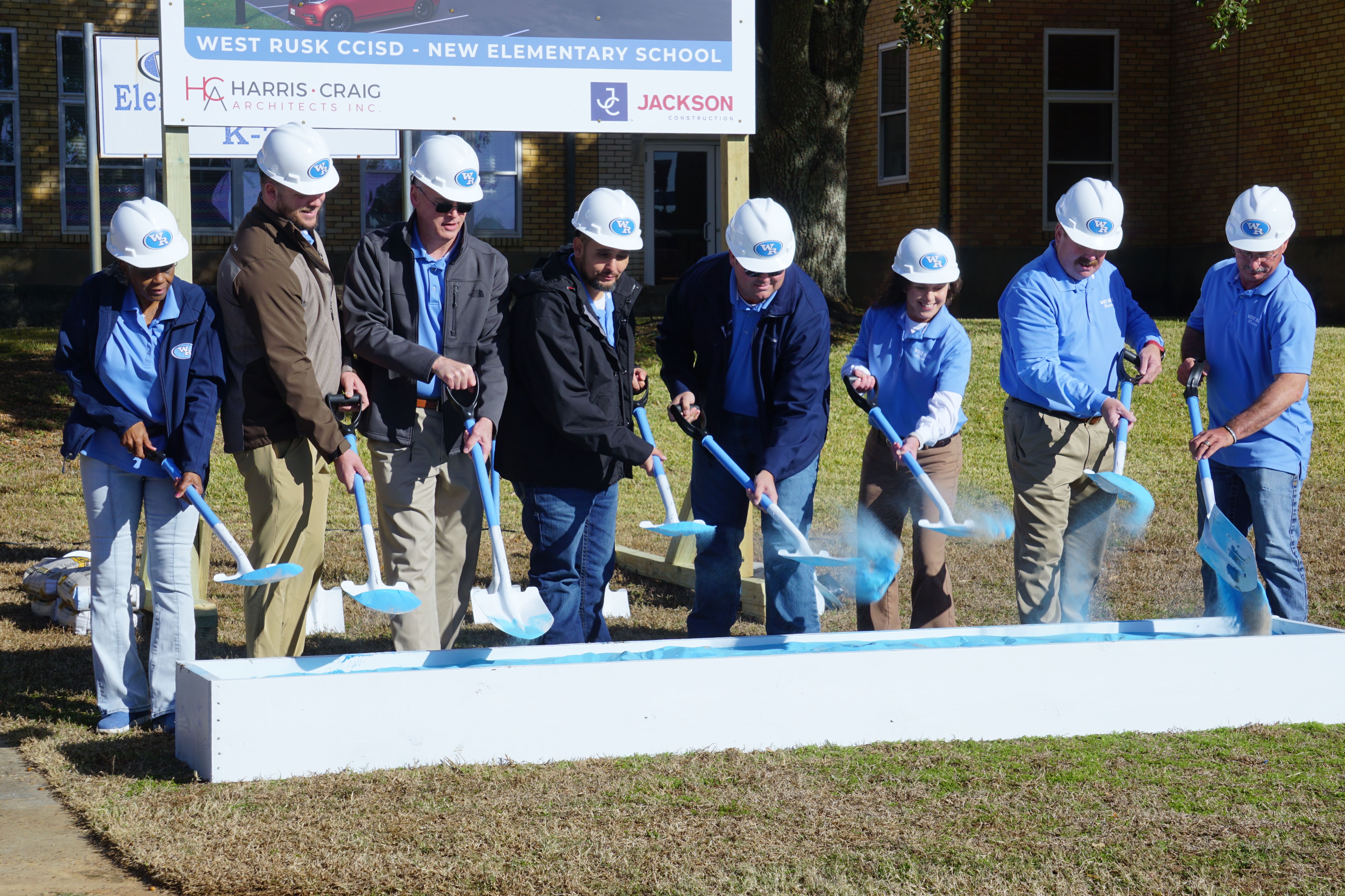 A group of people in hard hats and construction attire are gathered around a groundbreaking ceremony for a new elementary school, with a sign in the background indicating the school's name.