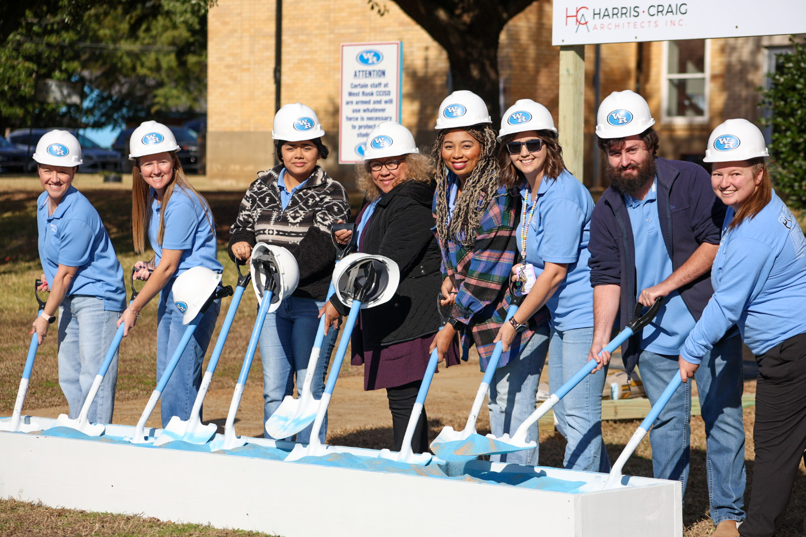 A group of people wearing hard hats and construction gear are standing in front of a building, holding shovels and appearing to be participating in a groundbreaking or construction event.