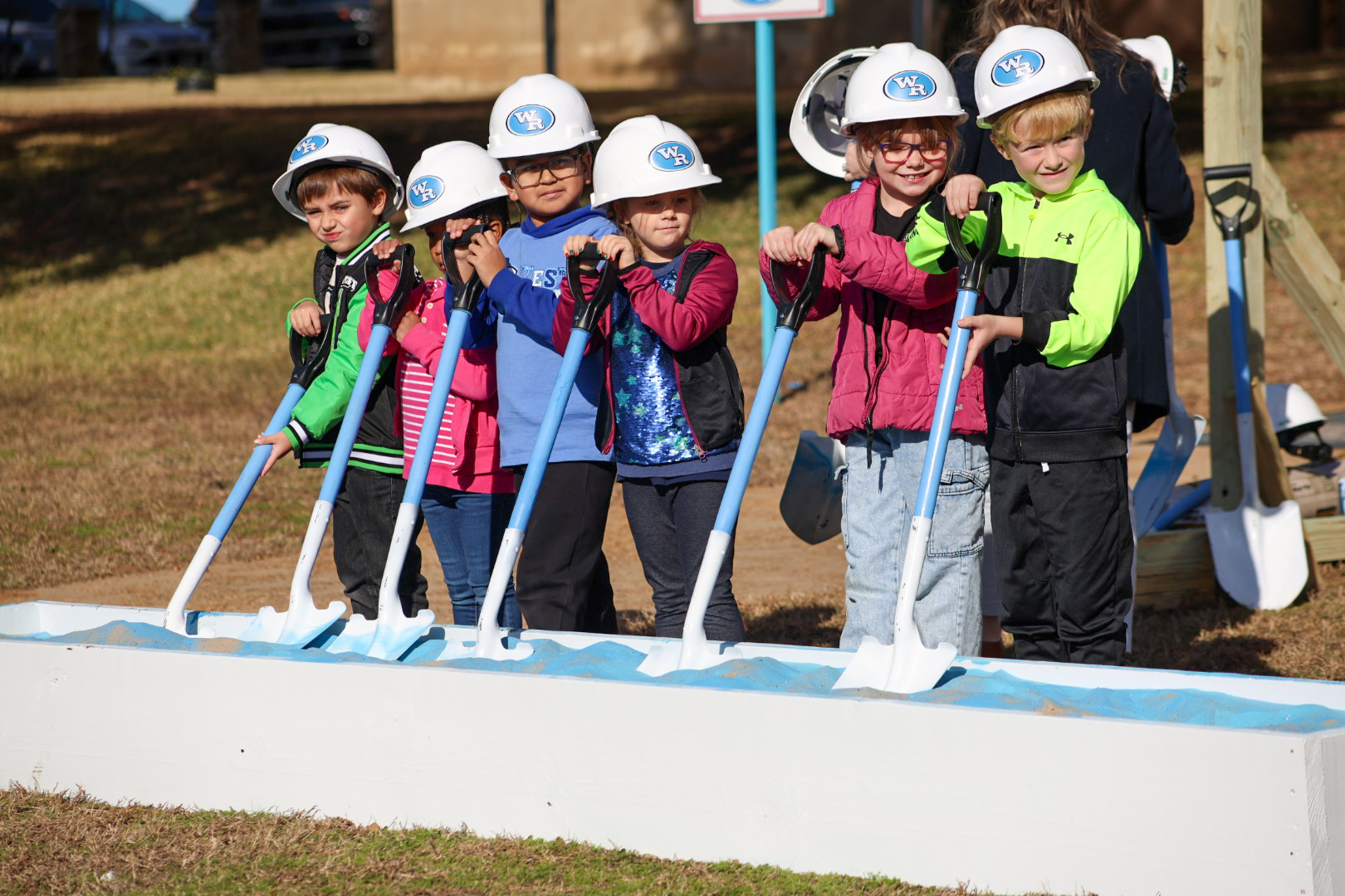 A group of children wearing hard hats and safety vests stand together on a construction site, with a building in the background.