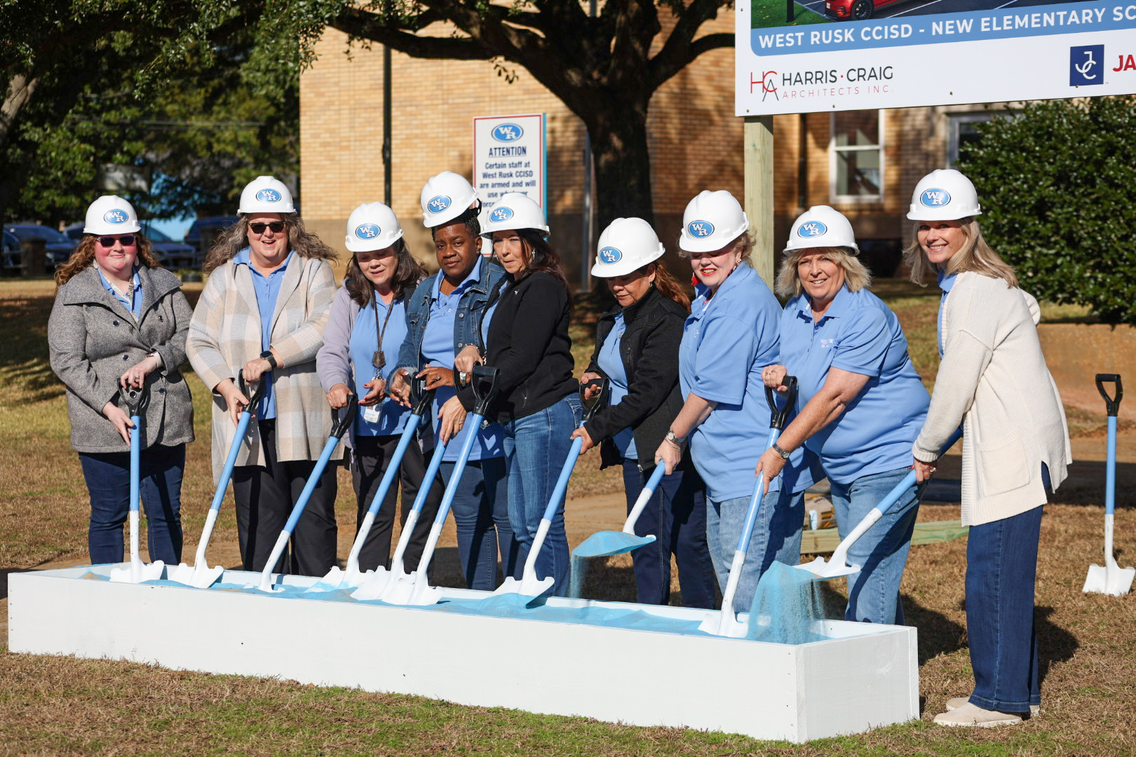 A group of people, wearing hard hats, stand together in front of a construction site, with a new elementary school building visible in the background.