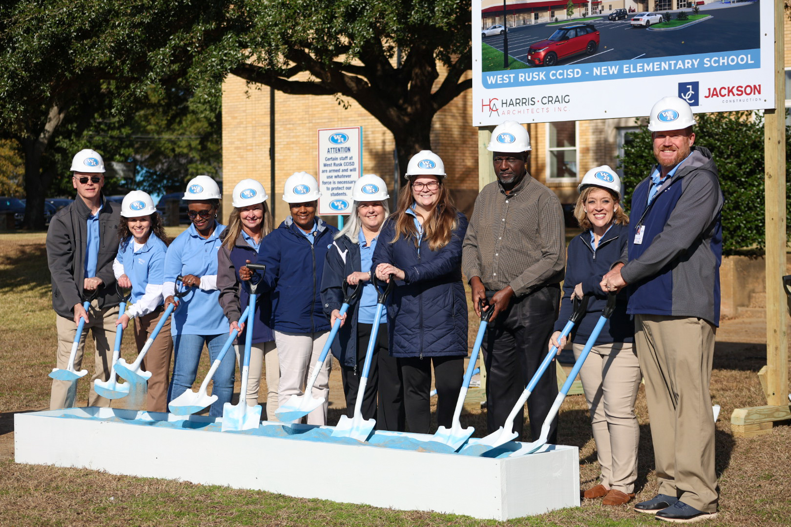 A group of people, likely school administrators or construction workers, are gathered in front of a school building, holding shovels and wearing hard hats, suggesting a groundbreaking or construction event.