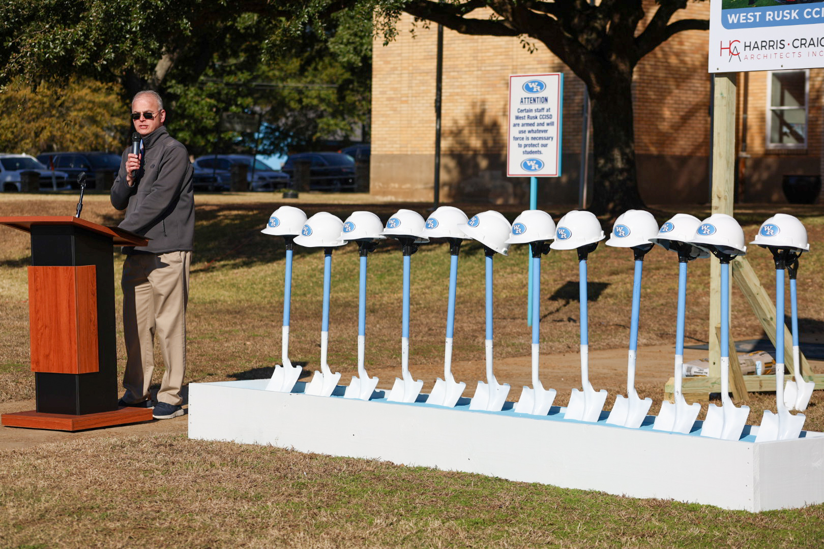 A man in a suit stands at a podium in front of a row of white hard hats on a grassy field, with a brick building and parked cars visible in the background.