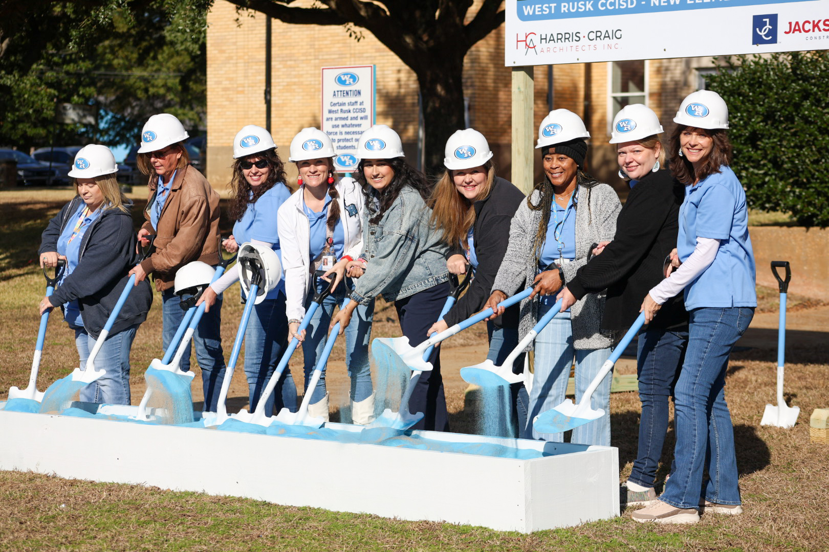 A group of people, likely construction workers or engineers, are gathered together holding shovels and wearing hard hats, standing in front of a construction site with a brick building in the background.