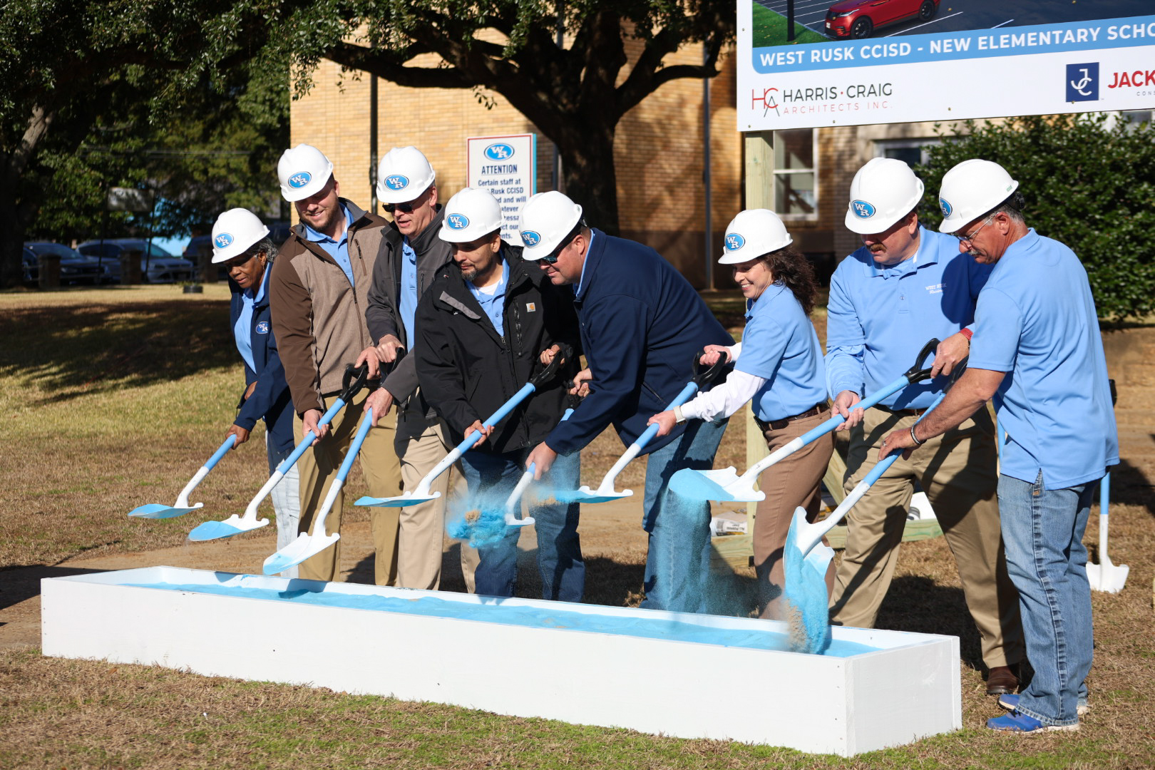 A group of people in hard hats and construction attire are gathered around a ceremonial groundbreaking event, with a new elementary school building visible in the background.