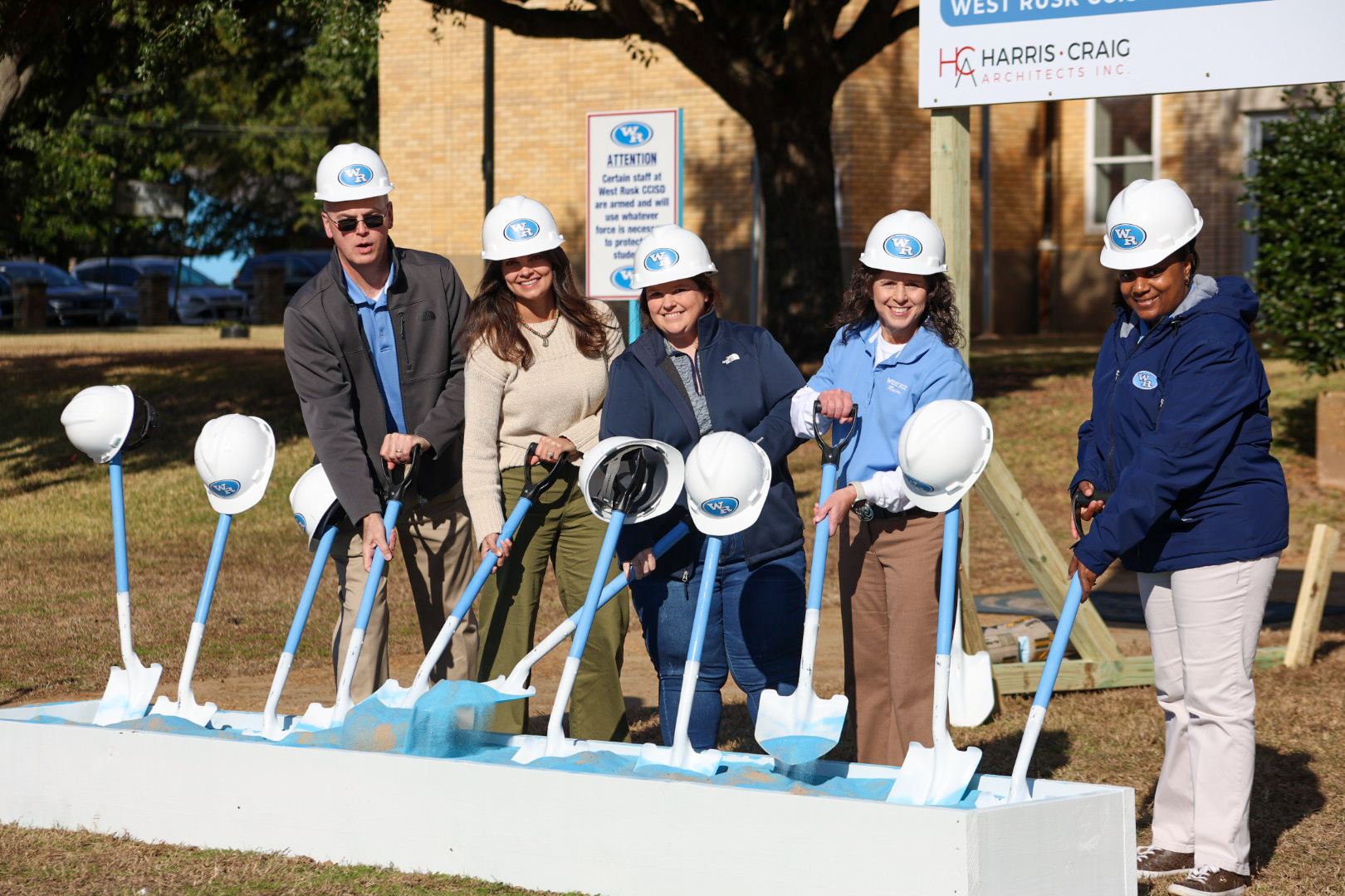 A group of people in hard hats and construction attire stand in front of a building, holding shovels and posing for a groundbreaking ceremony.