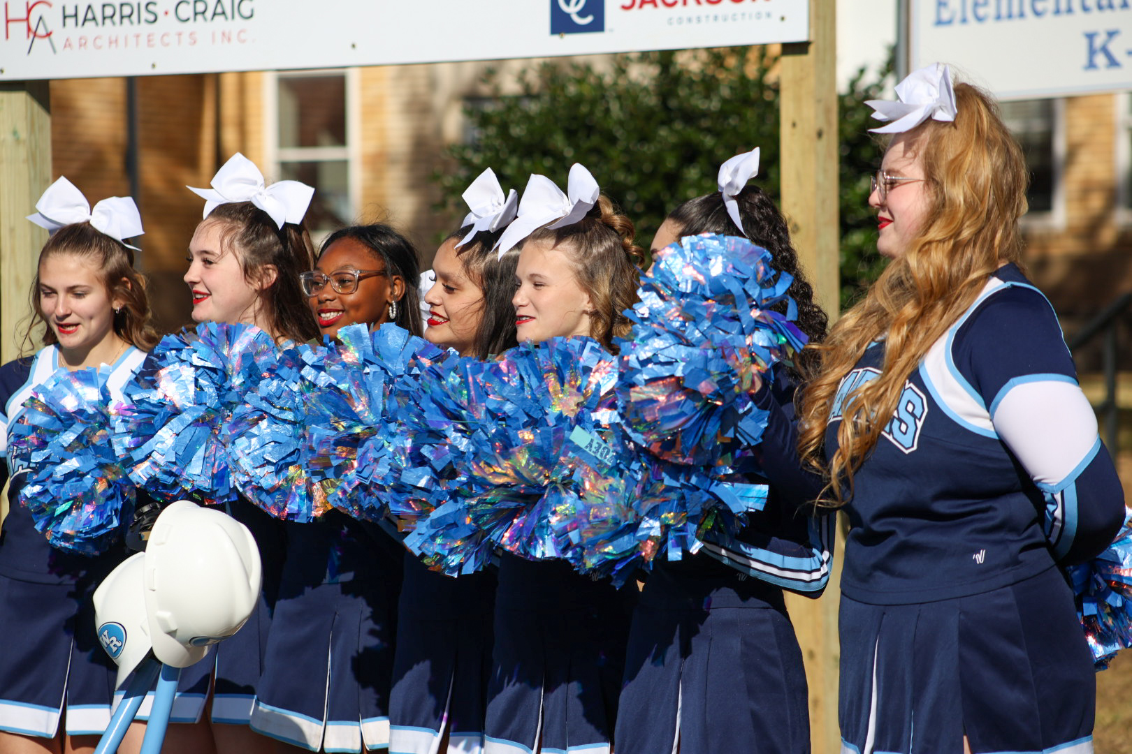 A group of cheerleaders in blue and white uniforms, with pom-poms, stand in front of a building with signage.