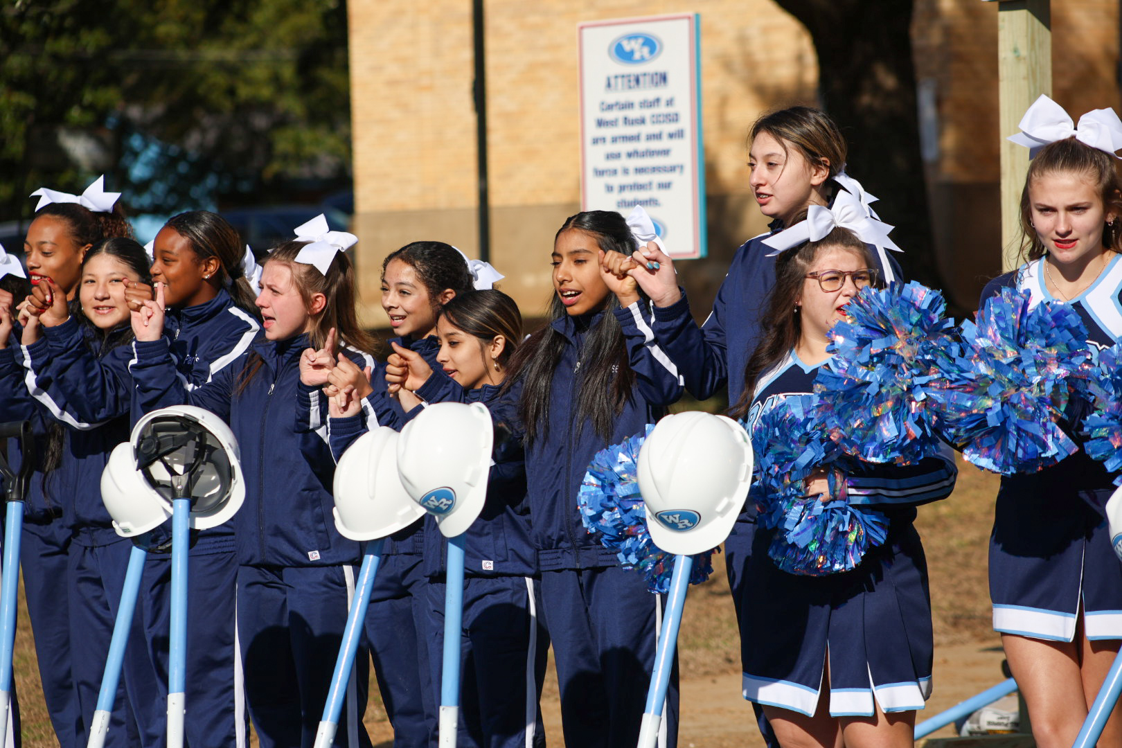 A group of young female cheerleaders in matching blue and white uniforms, holding pom-poms, stand in front of a building with a sign that appears to be a school or sports facility.