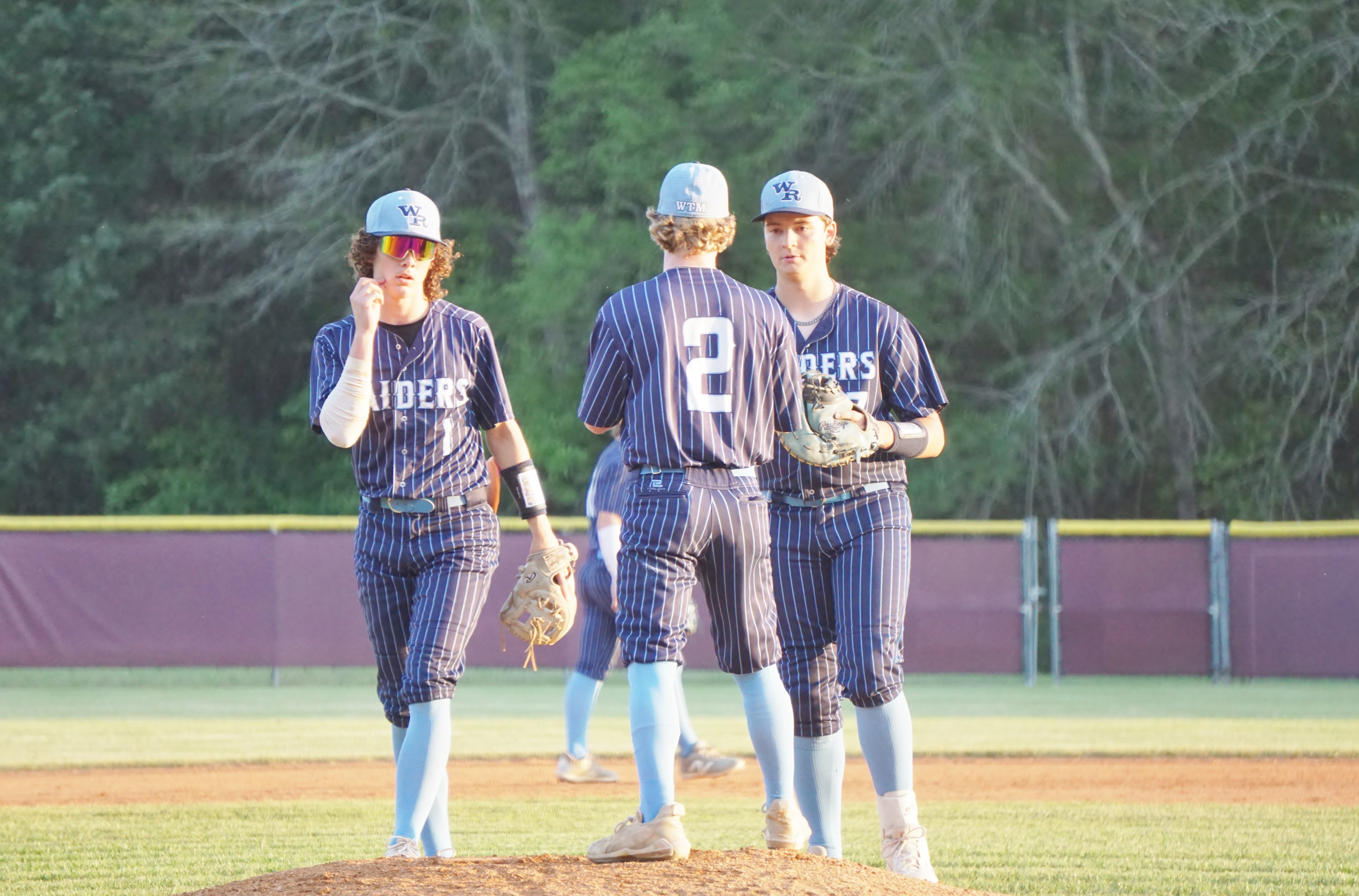 Three baseball players in uniform standing on a baseball field with a forested background.