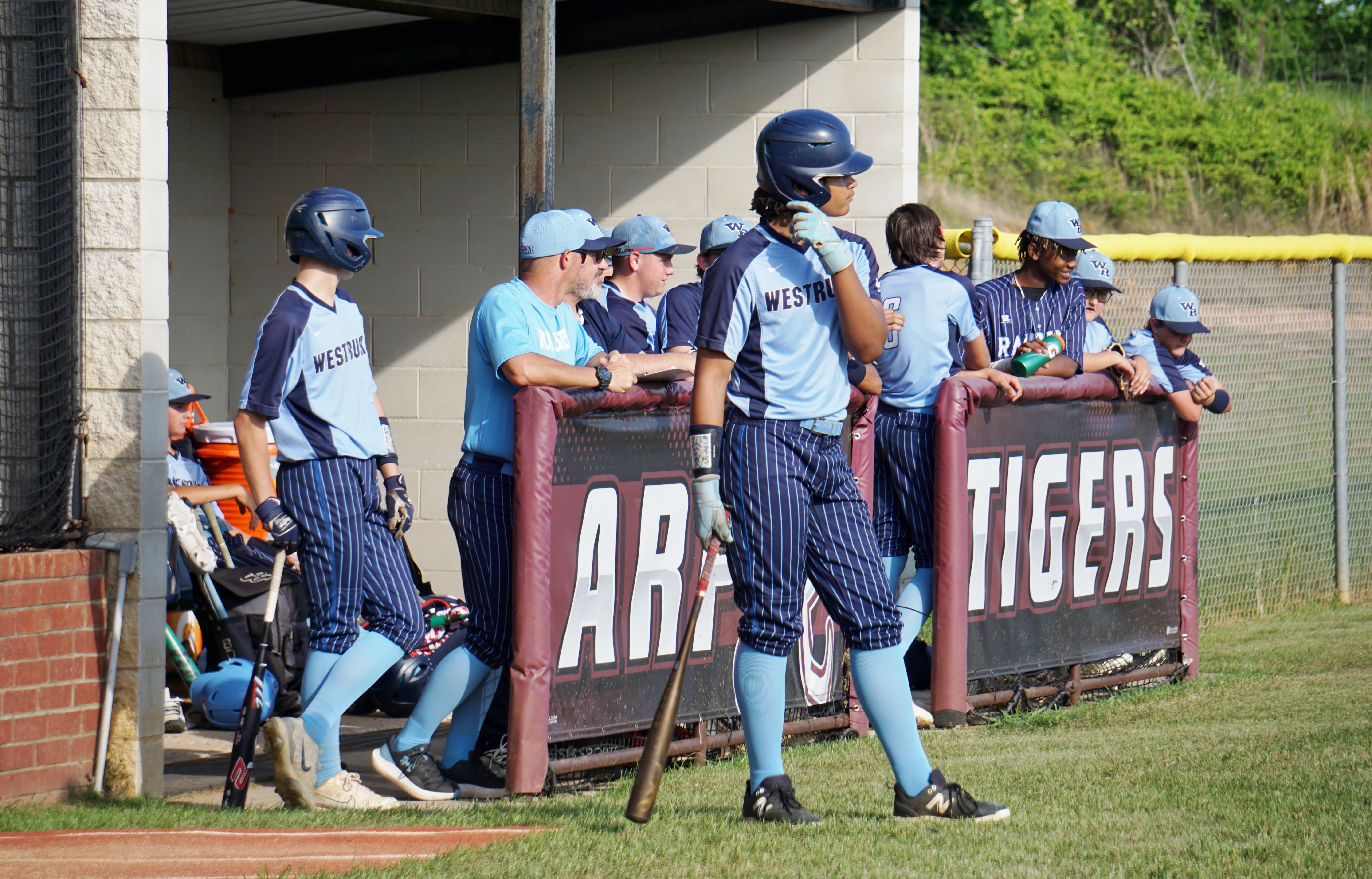 A group of baseball players in uniform standing near a dugout with the team name "TIGERS" displayed on the wall behind them.