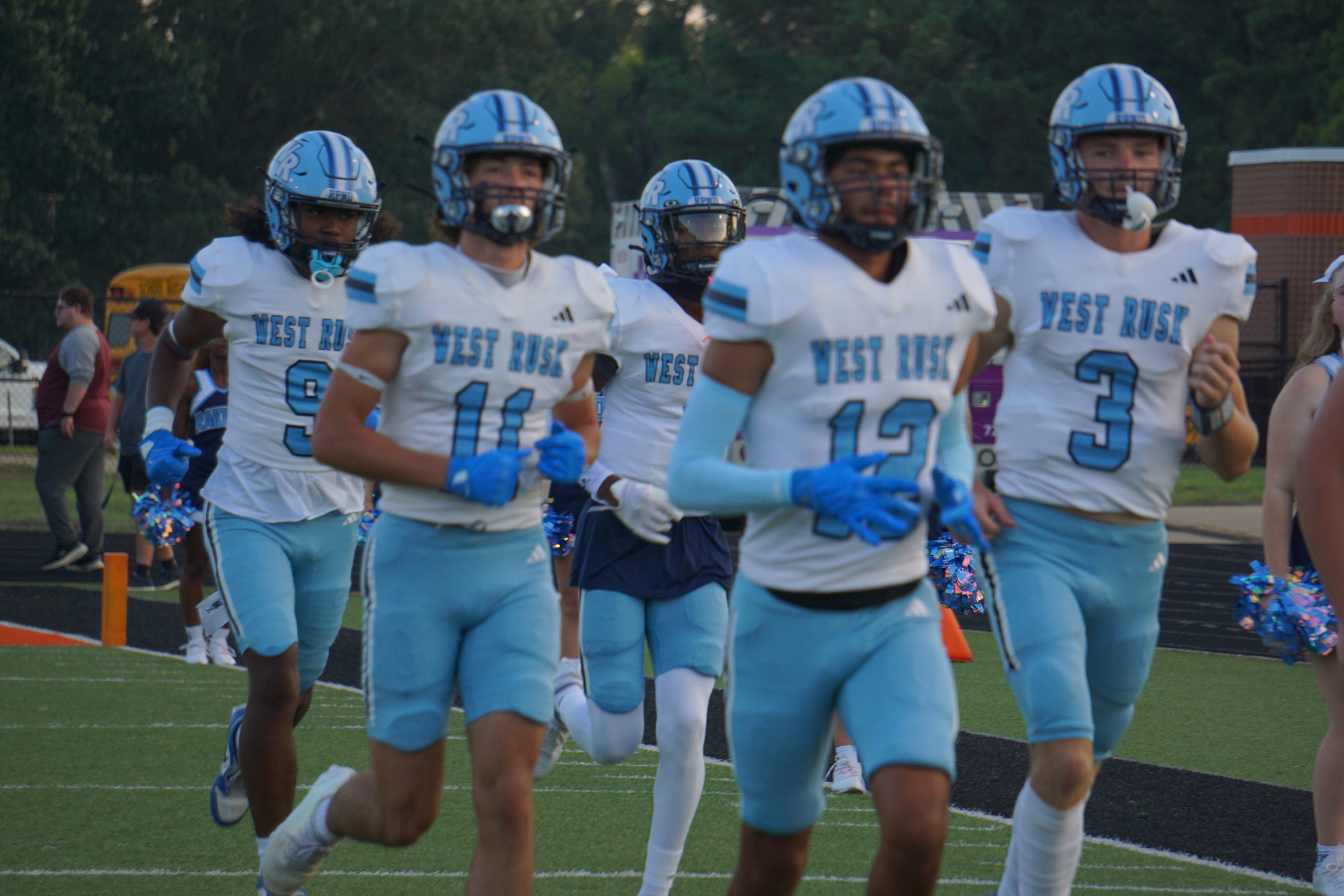 A group of football players in blue and white uniforms standing on a field, with a crowd visible in the background.