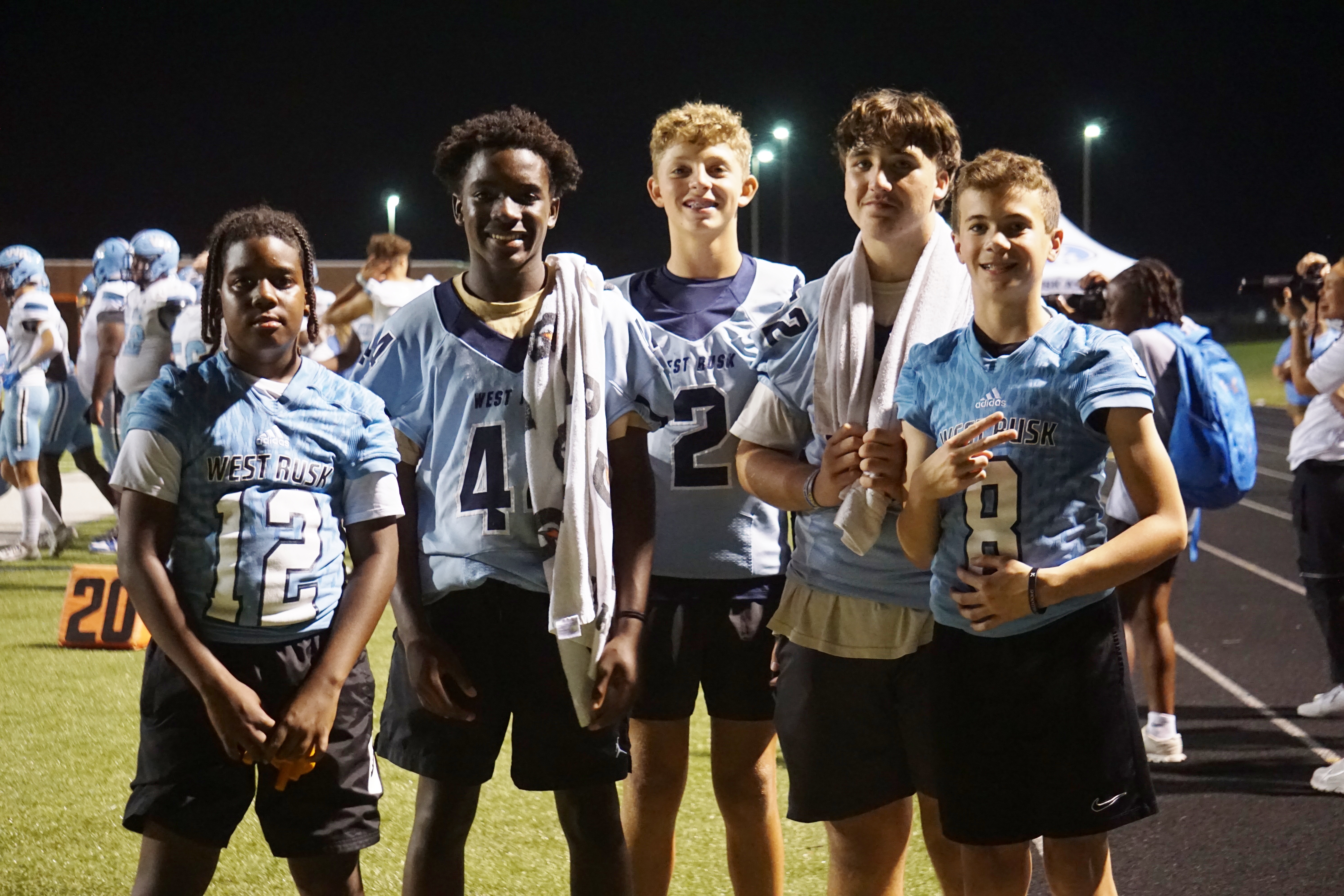 A group of young male athletes in sports uniforms standing together on a sports field at night, with stadium lights illuminating the scene.