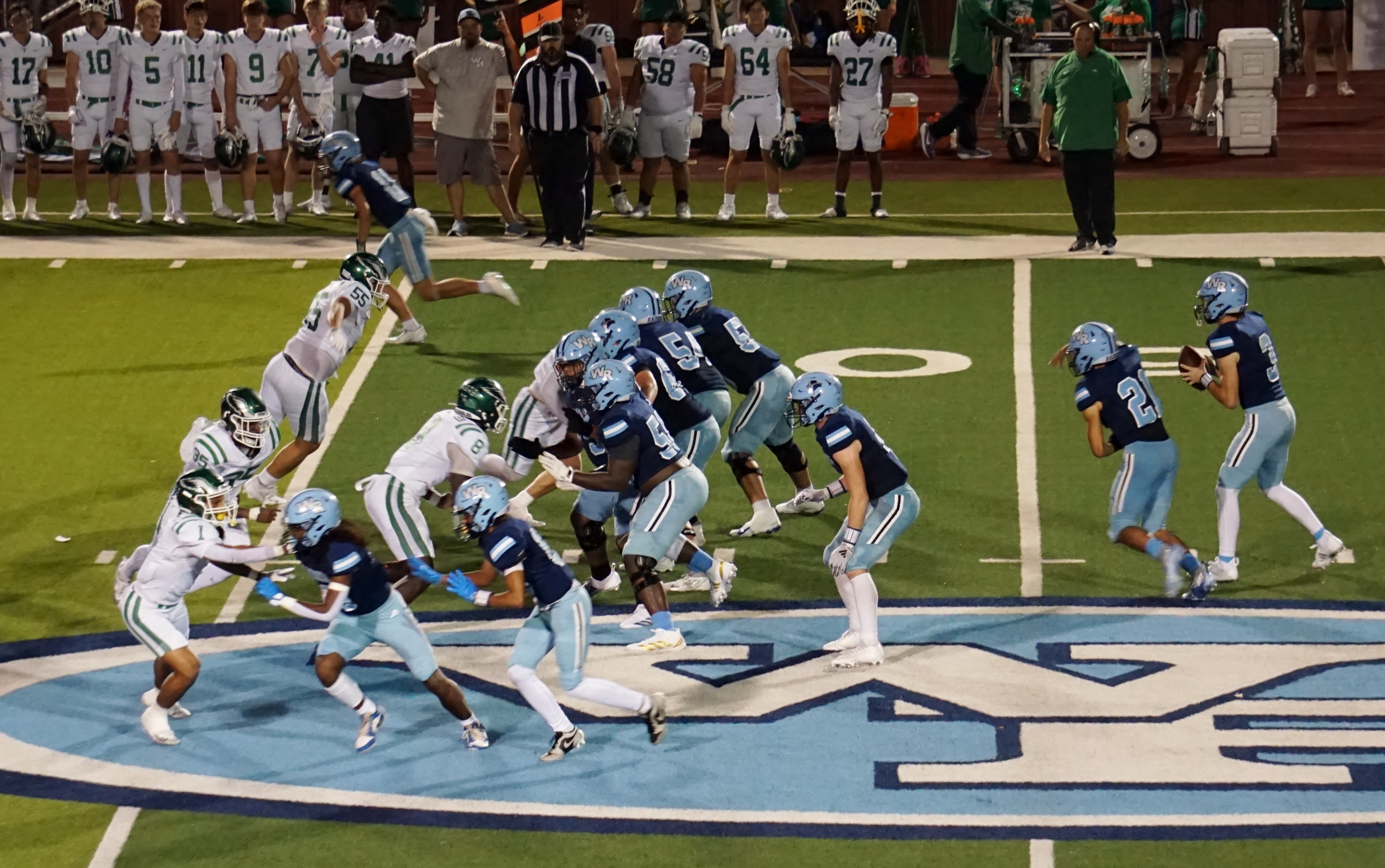 The image depicts a football game in progress, with players from both teams engaged in a play on the field. The players are wearing uniforms in different colors, and the background shows a stadium filled with spectators.