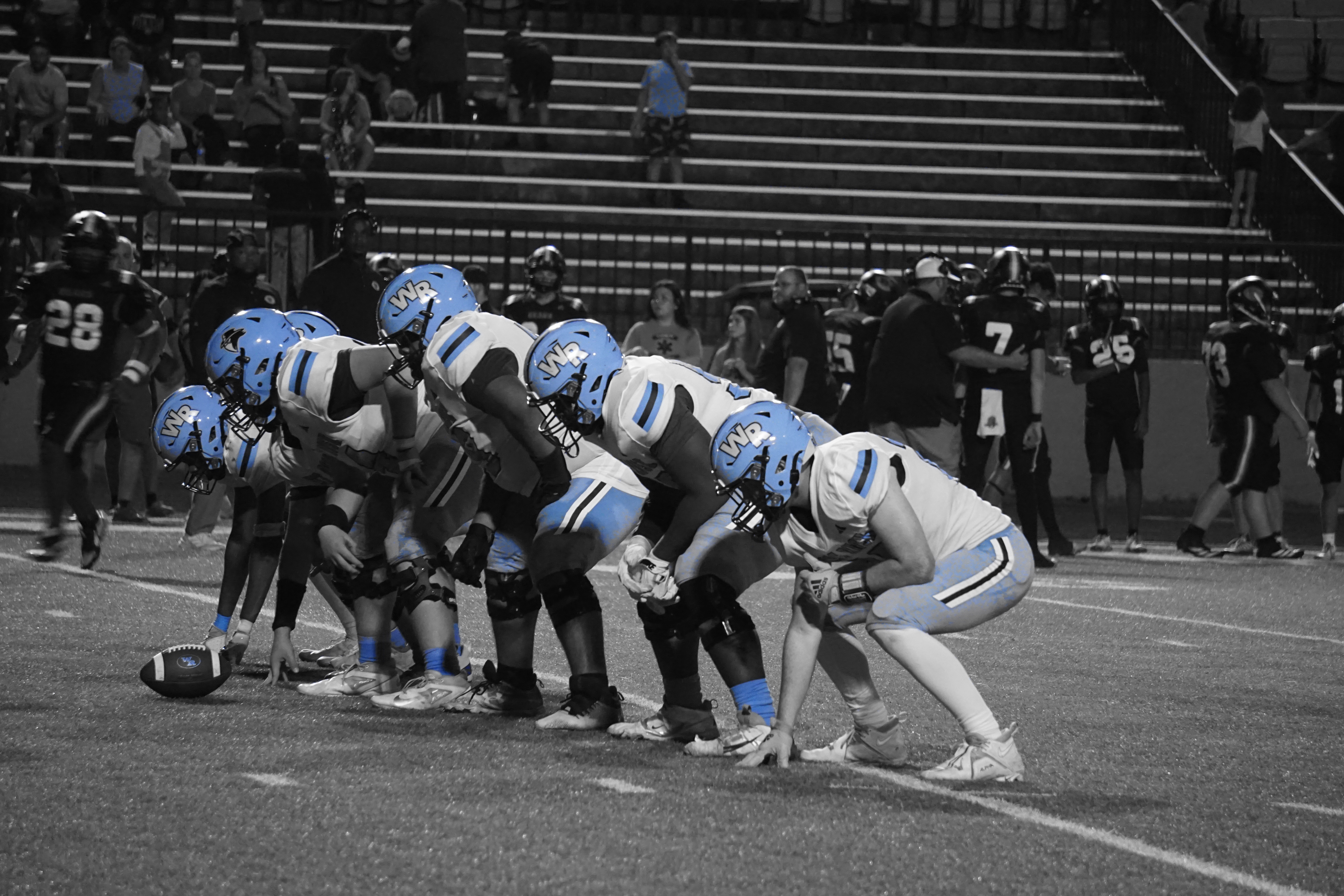 A group of football players in uniform are lined up on a field, with spectators visible in the background on stadium bleachers.