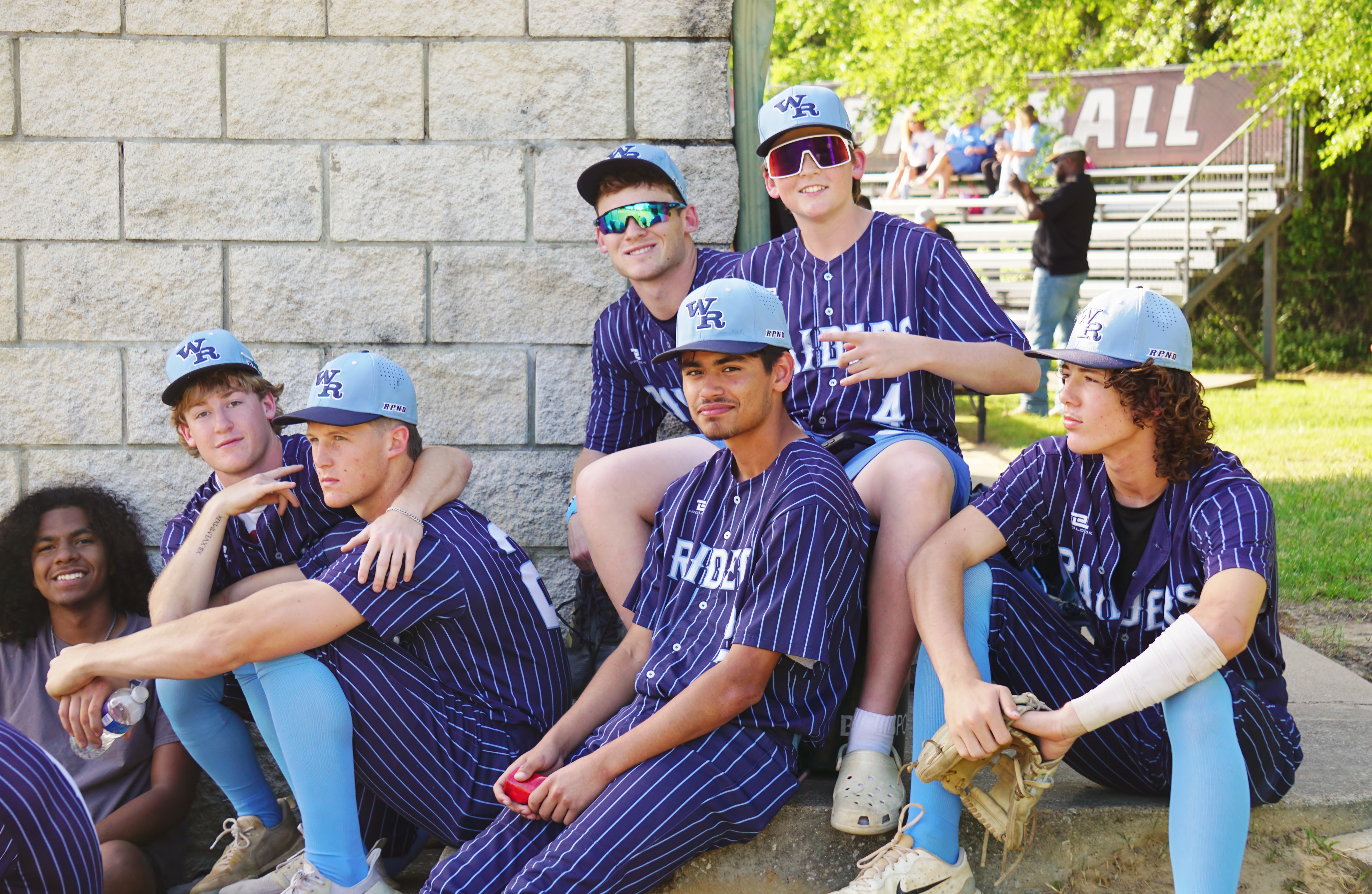 A group of young people wearing matching baseball uniforms stand together in front of a brick wall, with some individuals in the background visible.