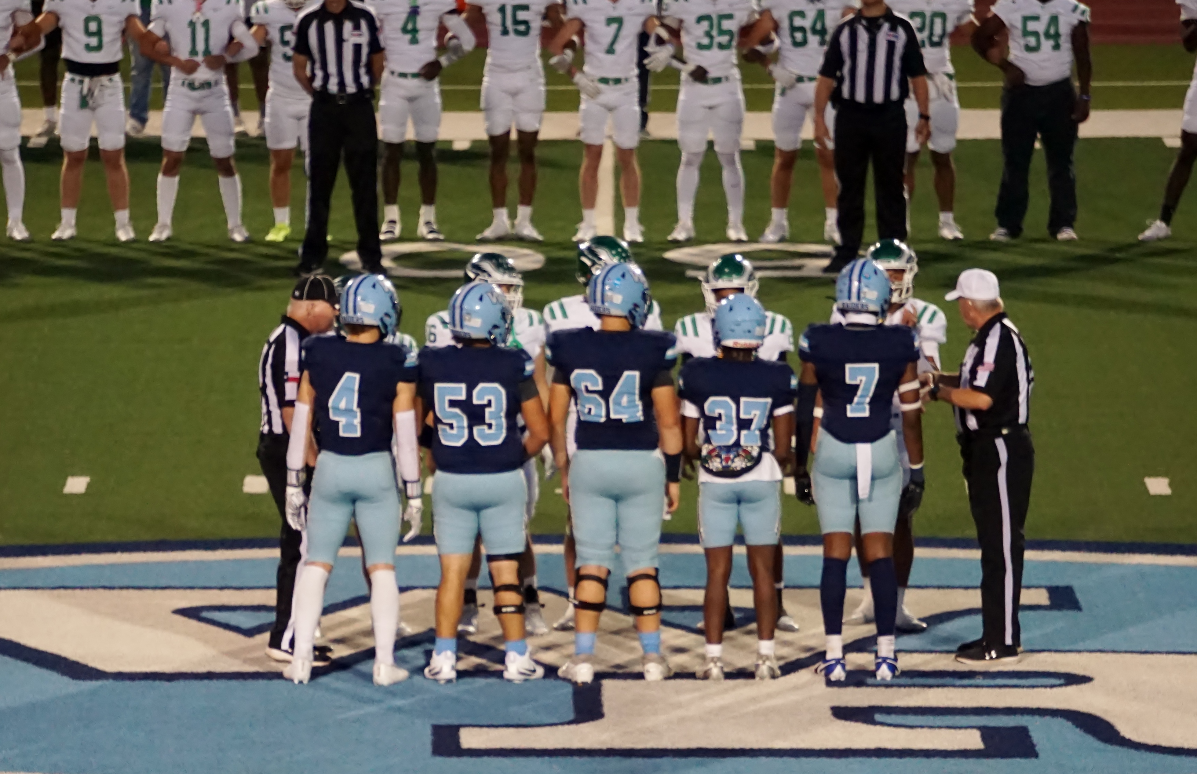 The image shows a group of football players, including referees, standing on a football field. The players are wearing uniforms in different colors, and the referees are wearing black and white striped shirts.
