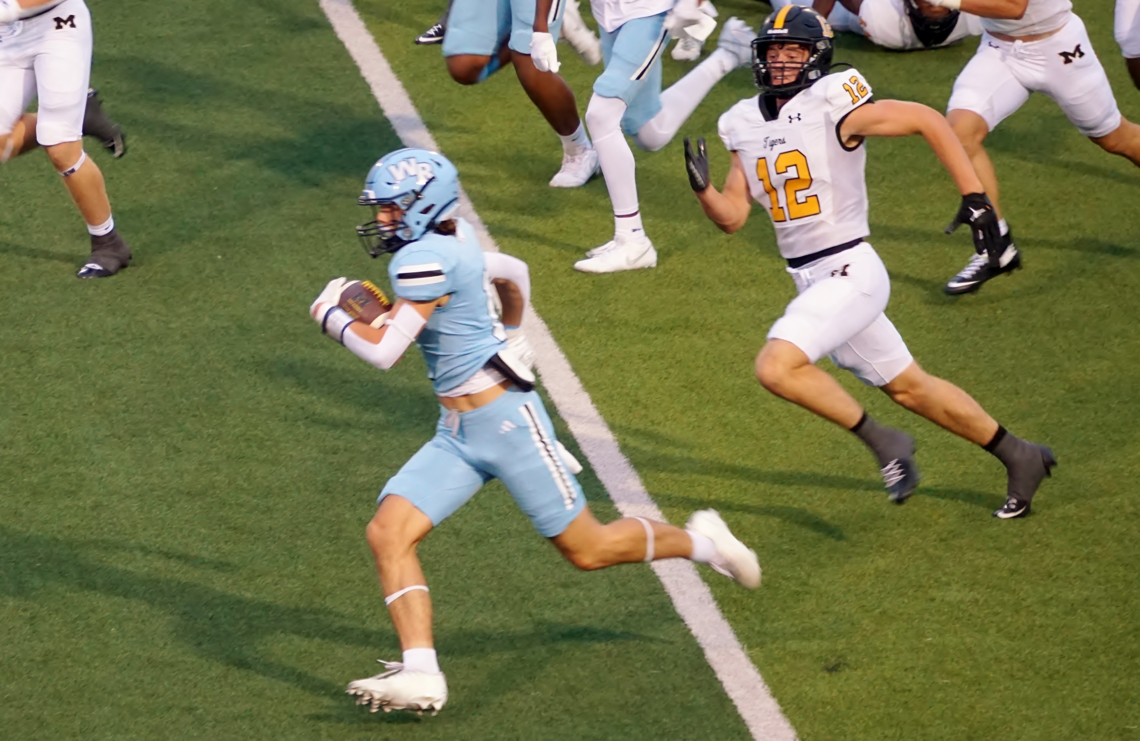 The image depicts a football game in progress, with players from both teams engaged in the action on the field. The player in the foreground is carrying the football, while other players can be seen in the background.