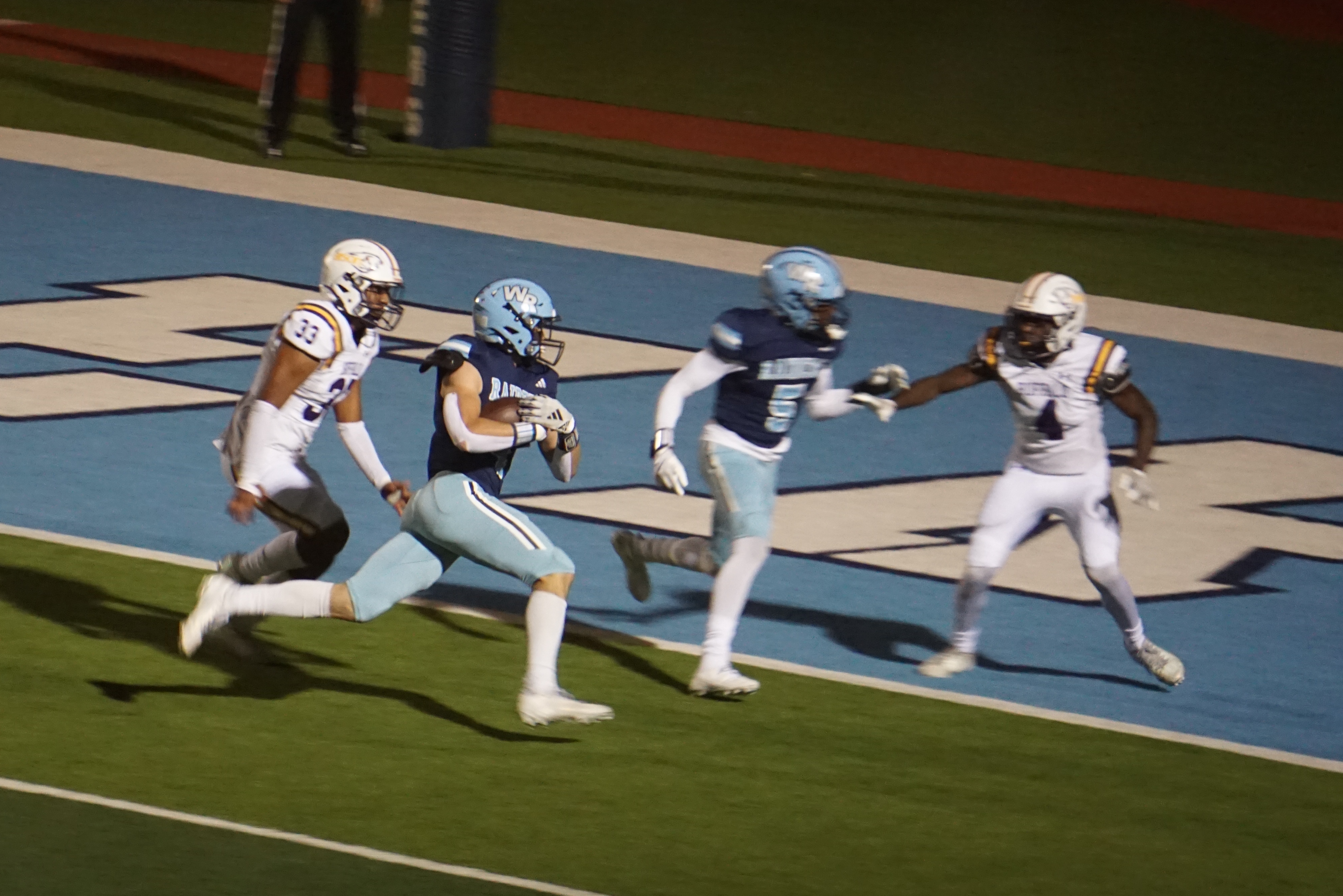 Four football players in uniform are running on a football field with a blue and white painted surface, surrounded by stadium seating in the background.