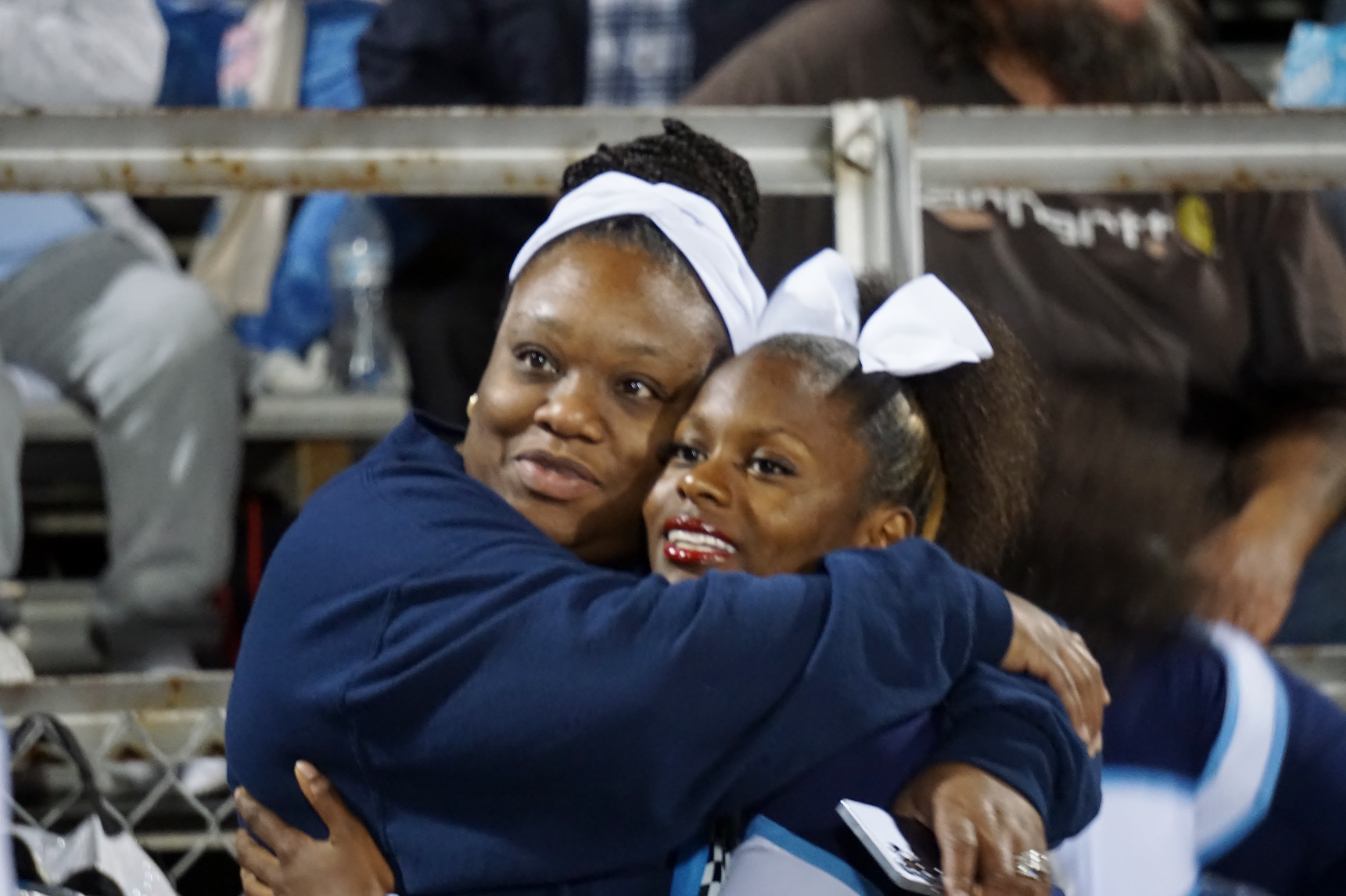 Two young African American girls in school uniforms are embracing each other in the foreground, with a blurred crowd of people visible in the background.