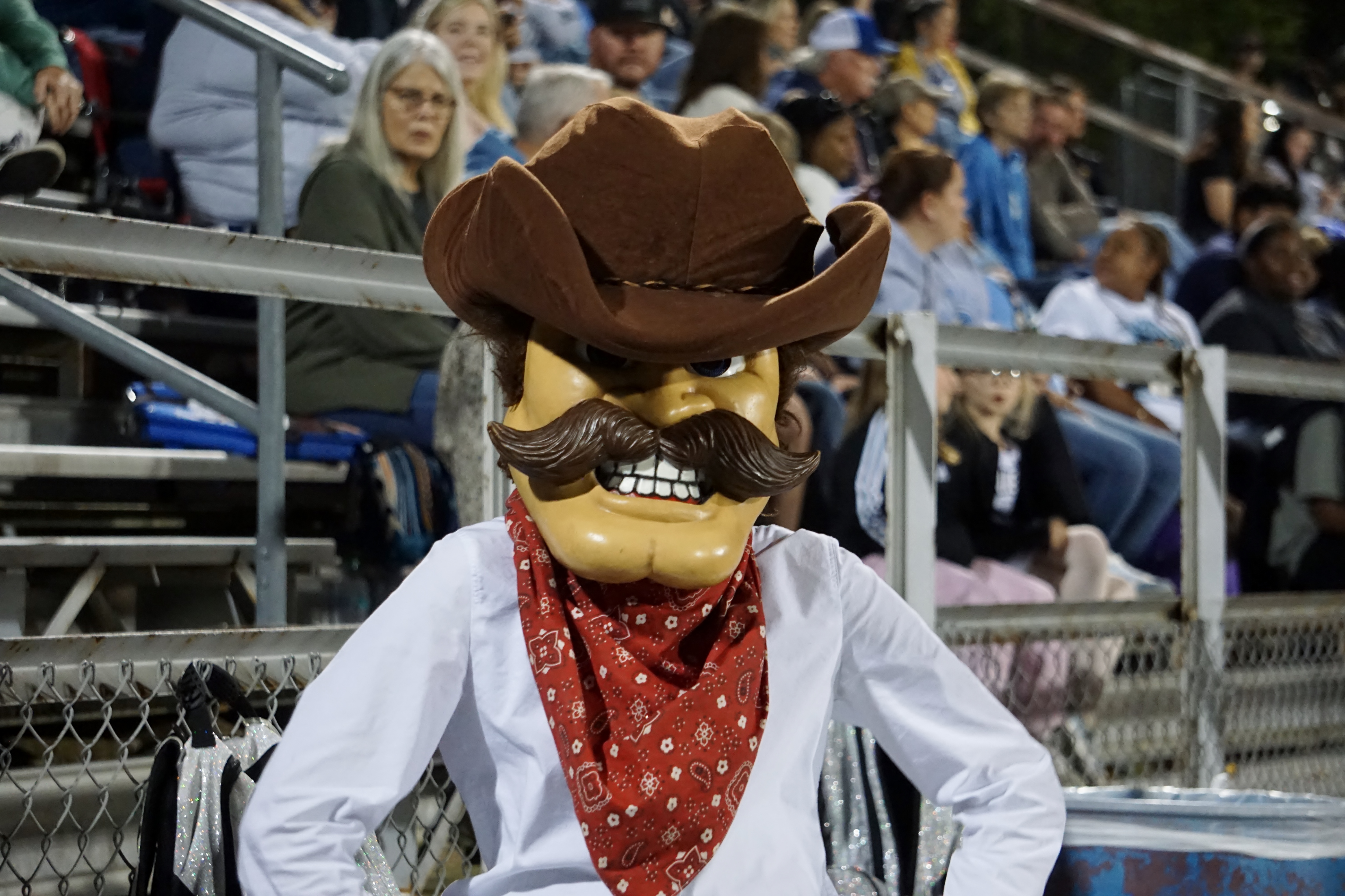 A person wearing a cowboy hat and a red bandana is standing in the foreground, with a crowd of people visible in the background.