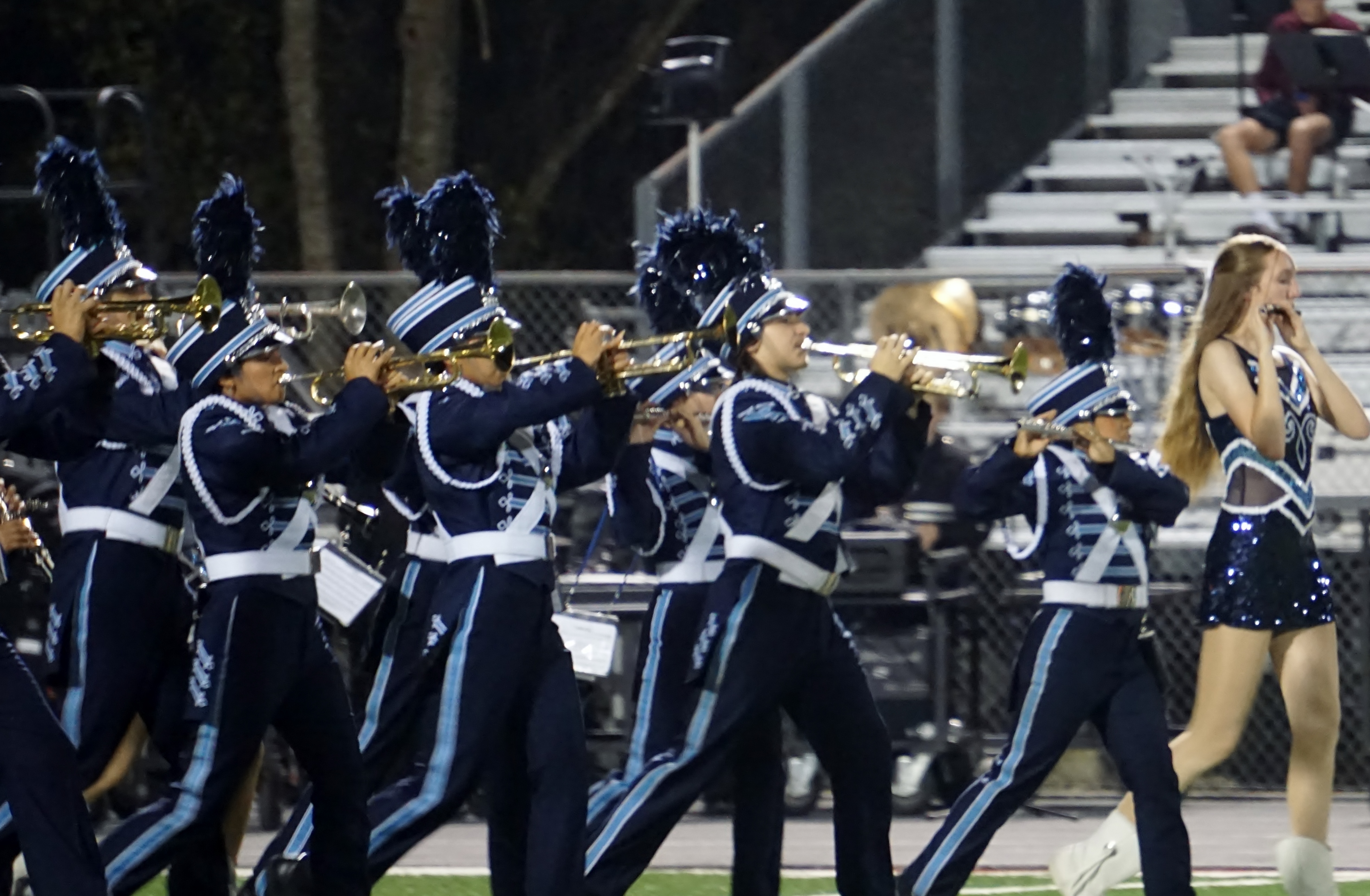 A marching band in uniform performing on a stage, with spectators visible in the background.