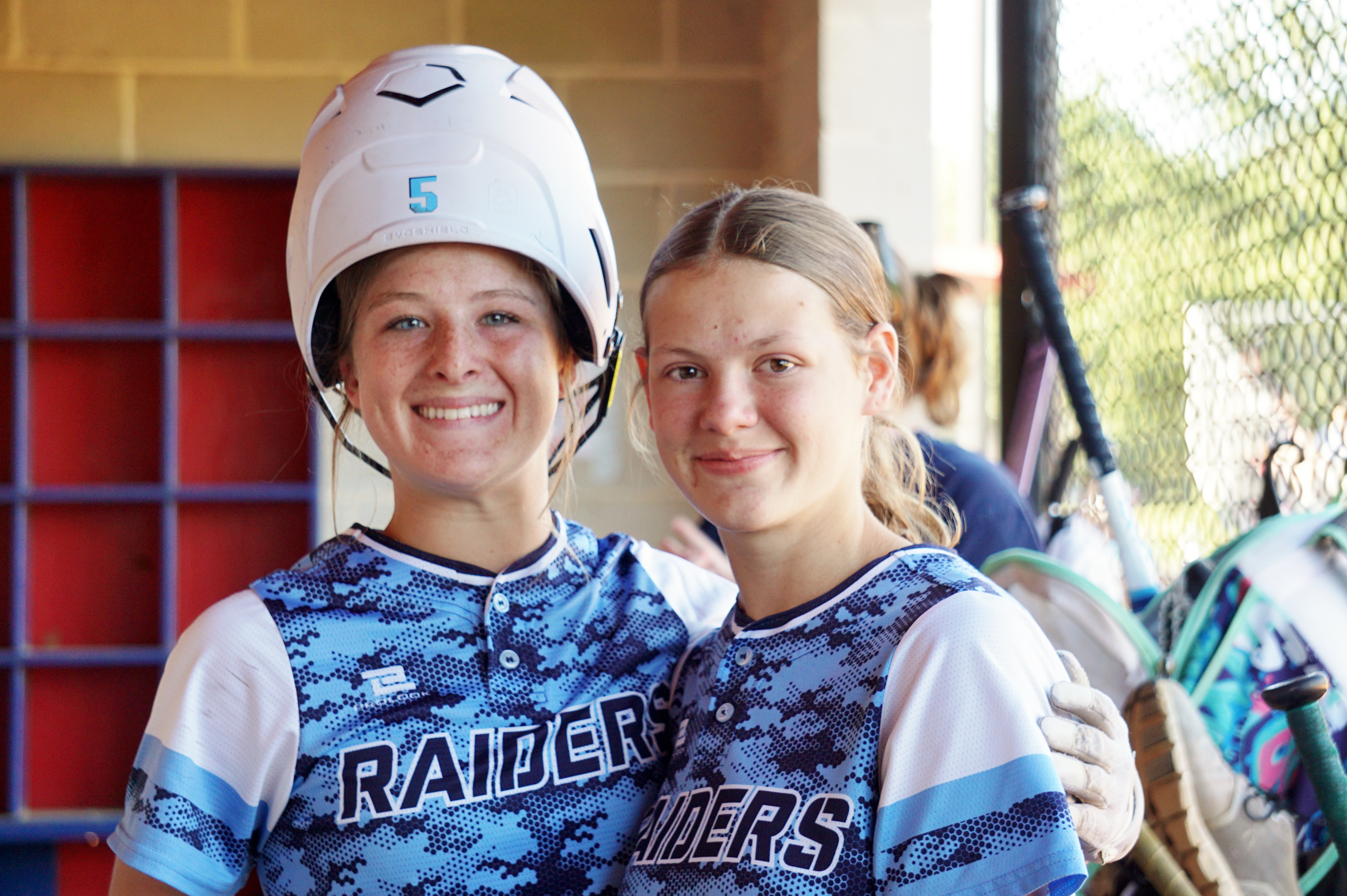 Two smiling young women wearing sports uniforms and helmets, standing in front of a red and white background.