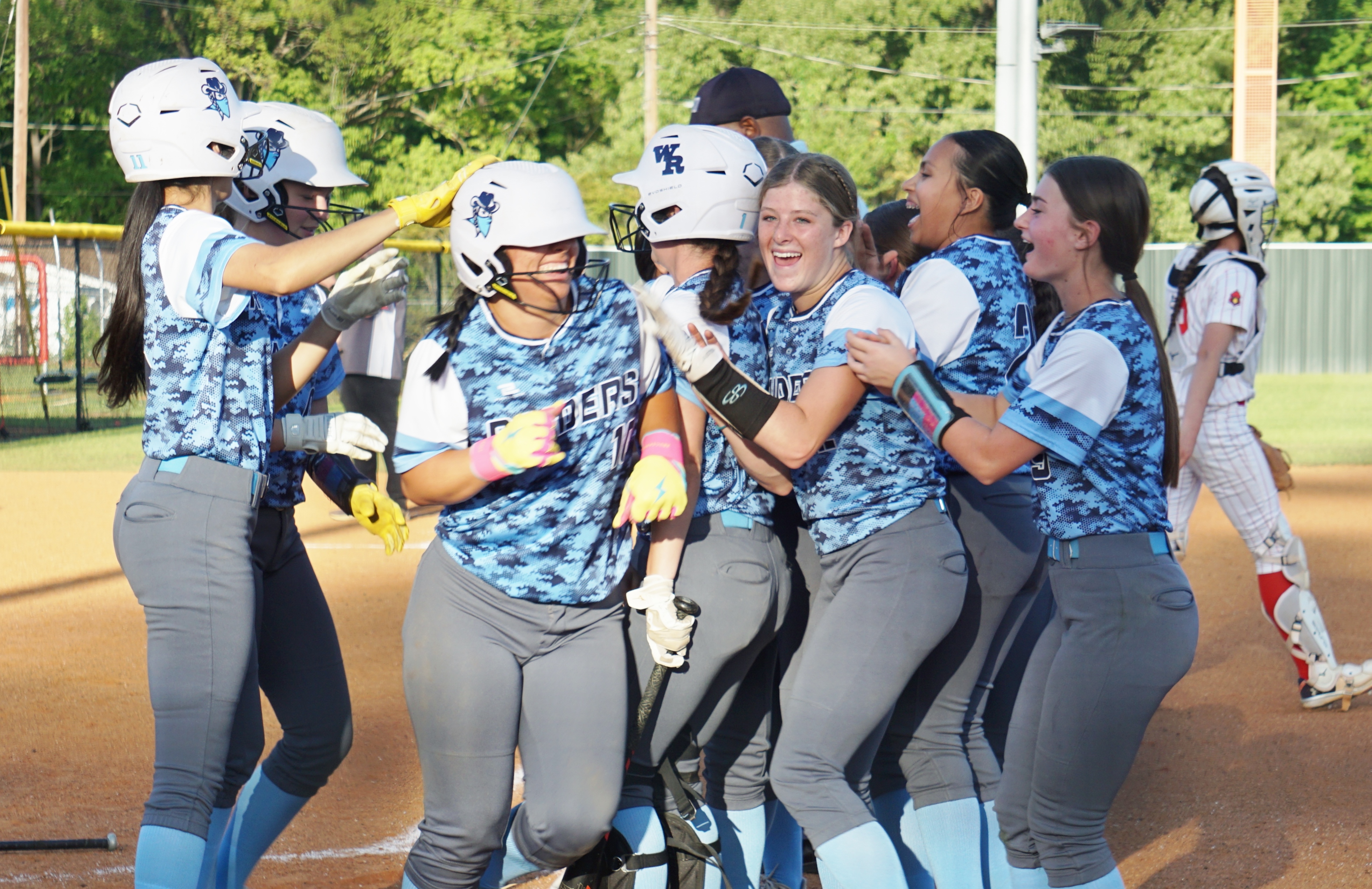 A group of young women in matching uniforms are gathered together, laughing and celebrating on a baseball field surrounded by lush greenery.