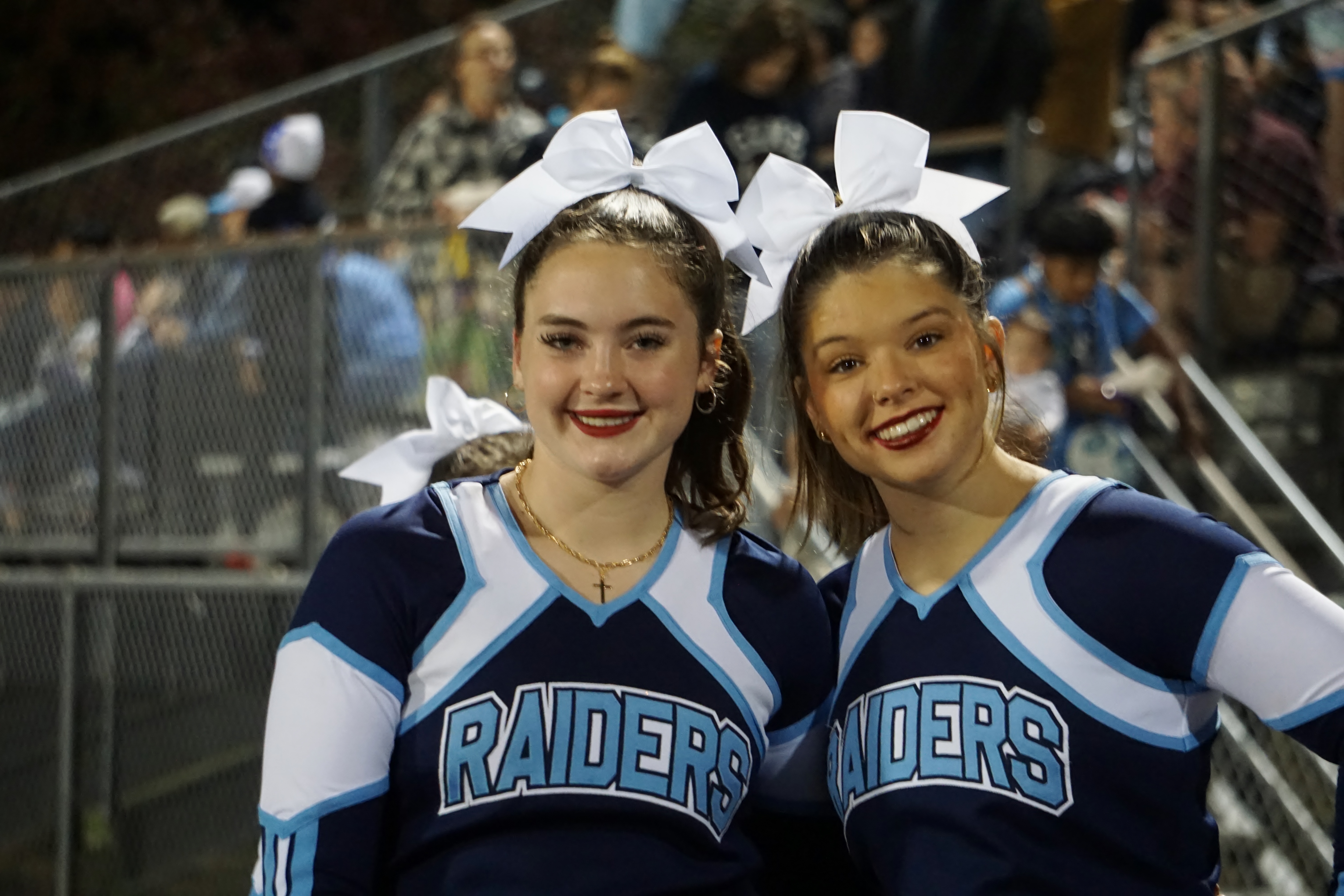 Two young women wearing cheerleader uniforms with the word "Raiders" on them are smiling and posing together in front of a crowd at what appears to be a sporting event.
