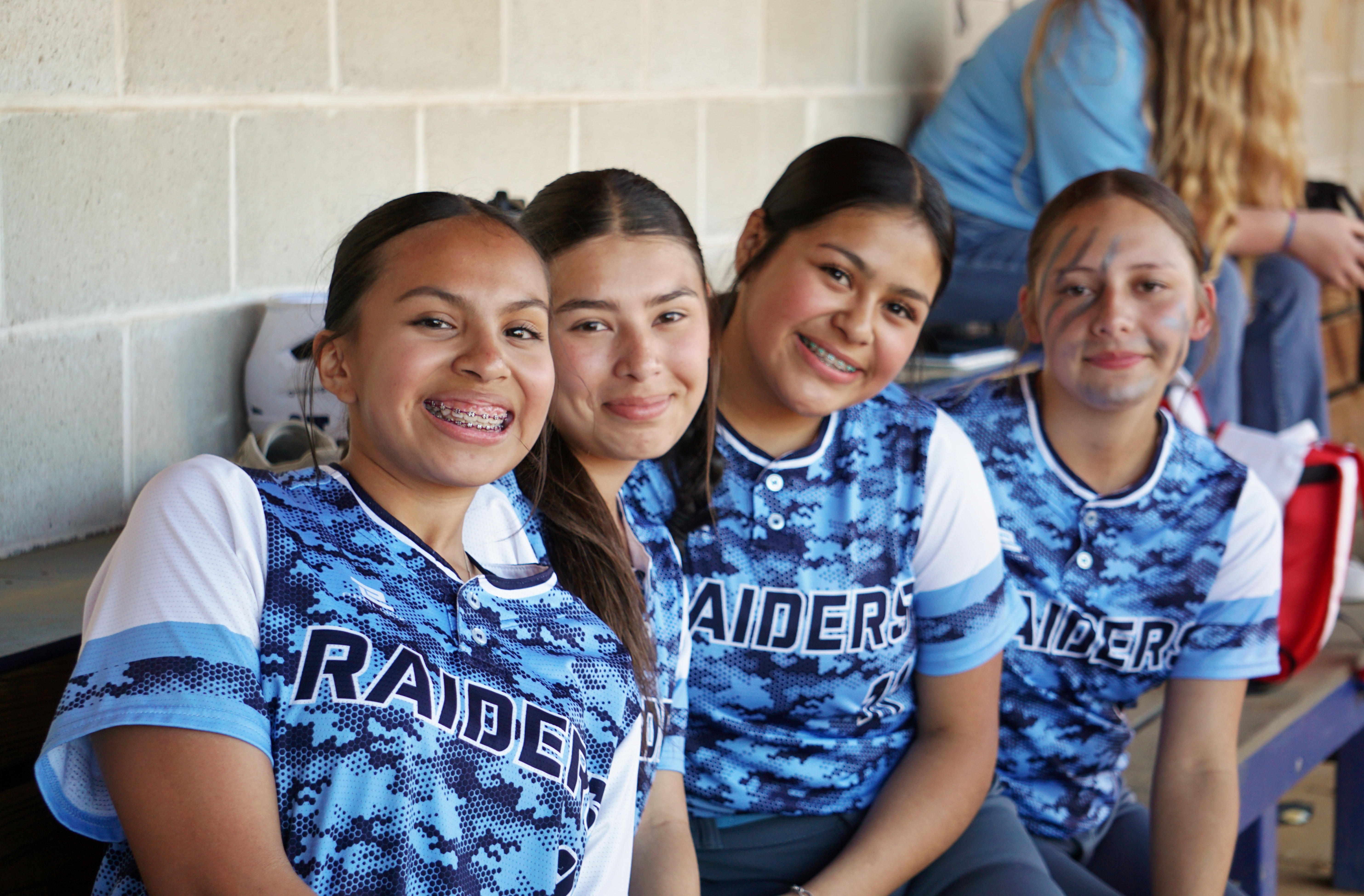 A group of young women wearing matching blue and white sports jerseys with the word "Raiders" printed on them, posing together in what appears to be a locker room or sports facility setting.