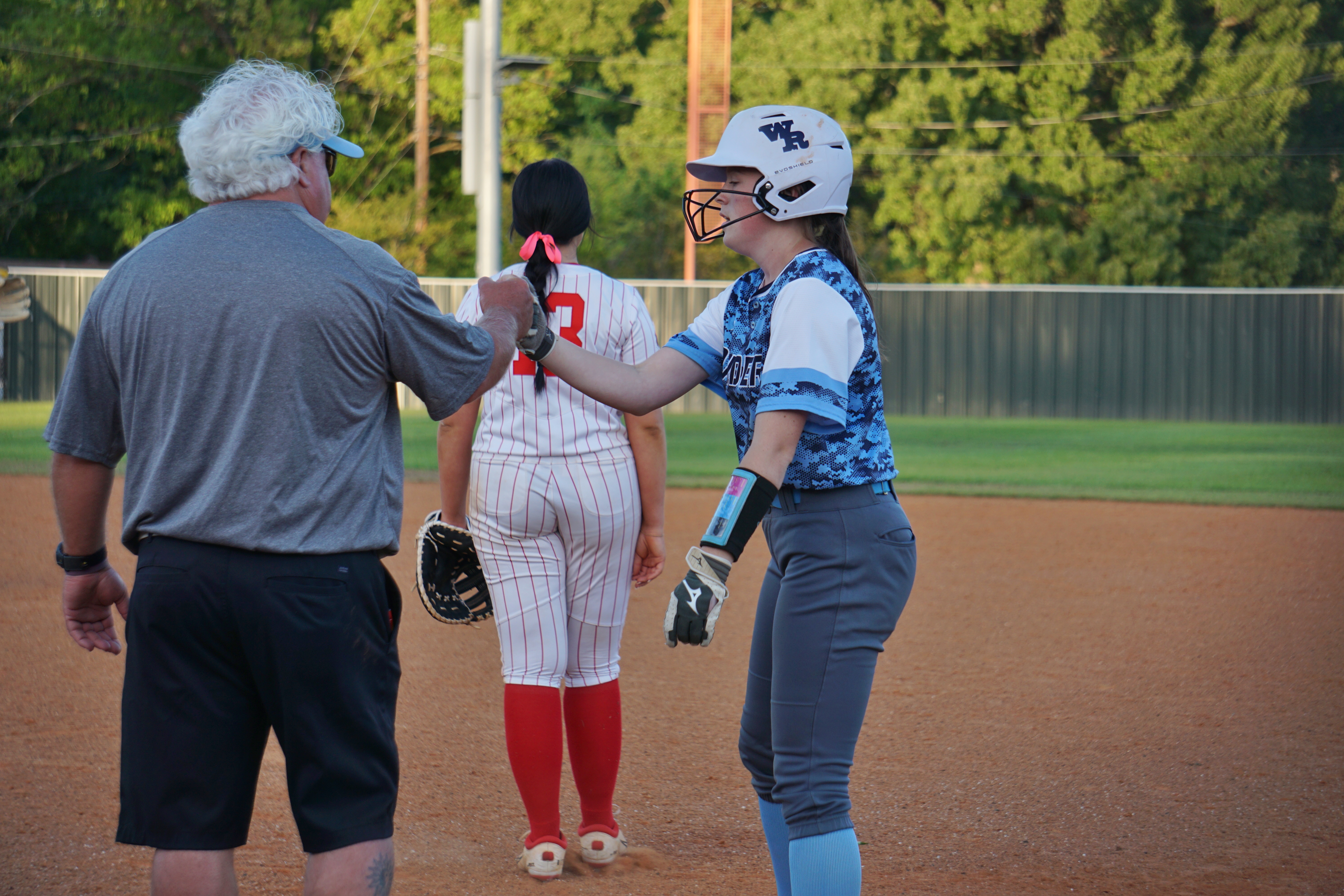The image shows two people, an older man and a younger woman, standing on a baseball field with a fence and trees in the background.