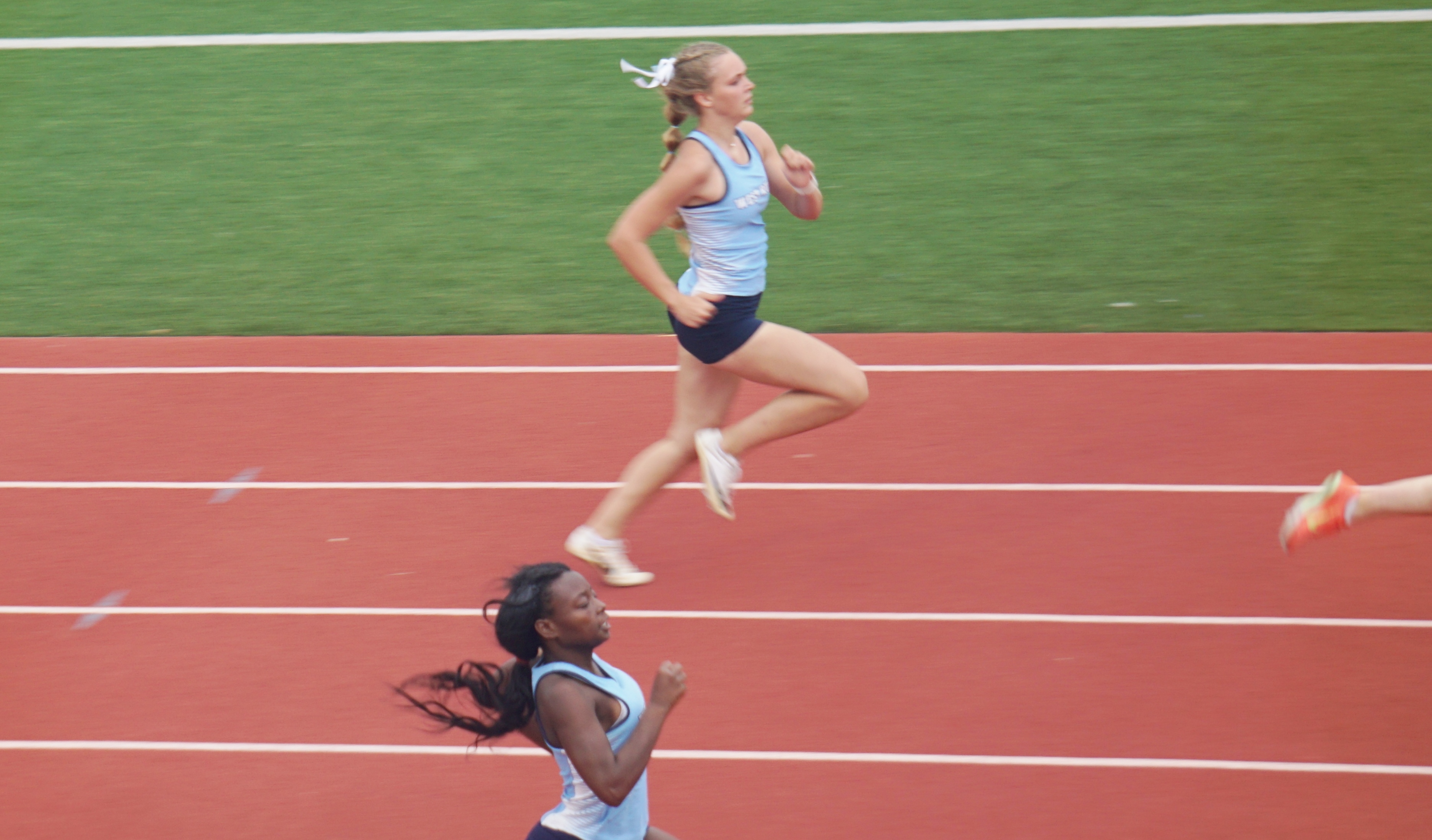 The image shows two female athletes competing on a running track, with one runner in the foreground and another in the background.