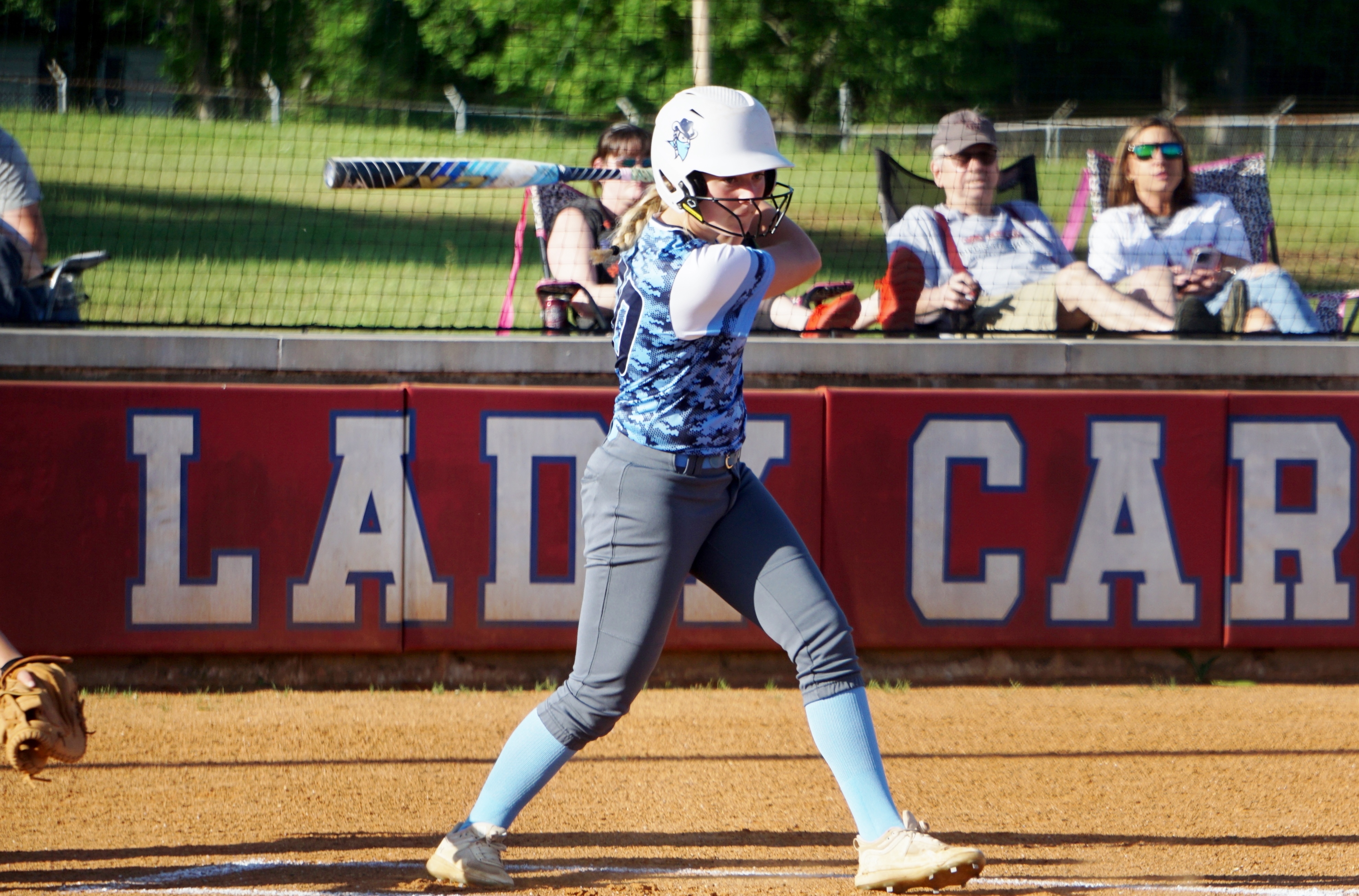 A young woman in a softball uniform stands on a baseball field, with a group of people sitting in the background.