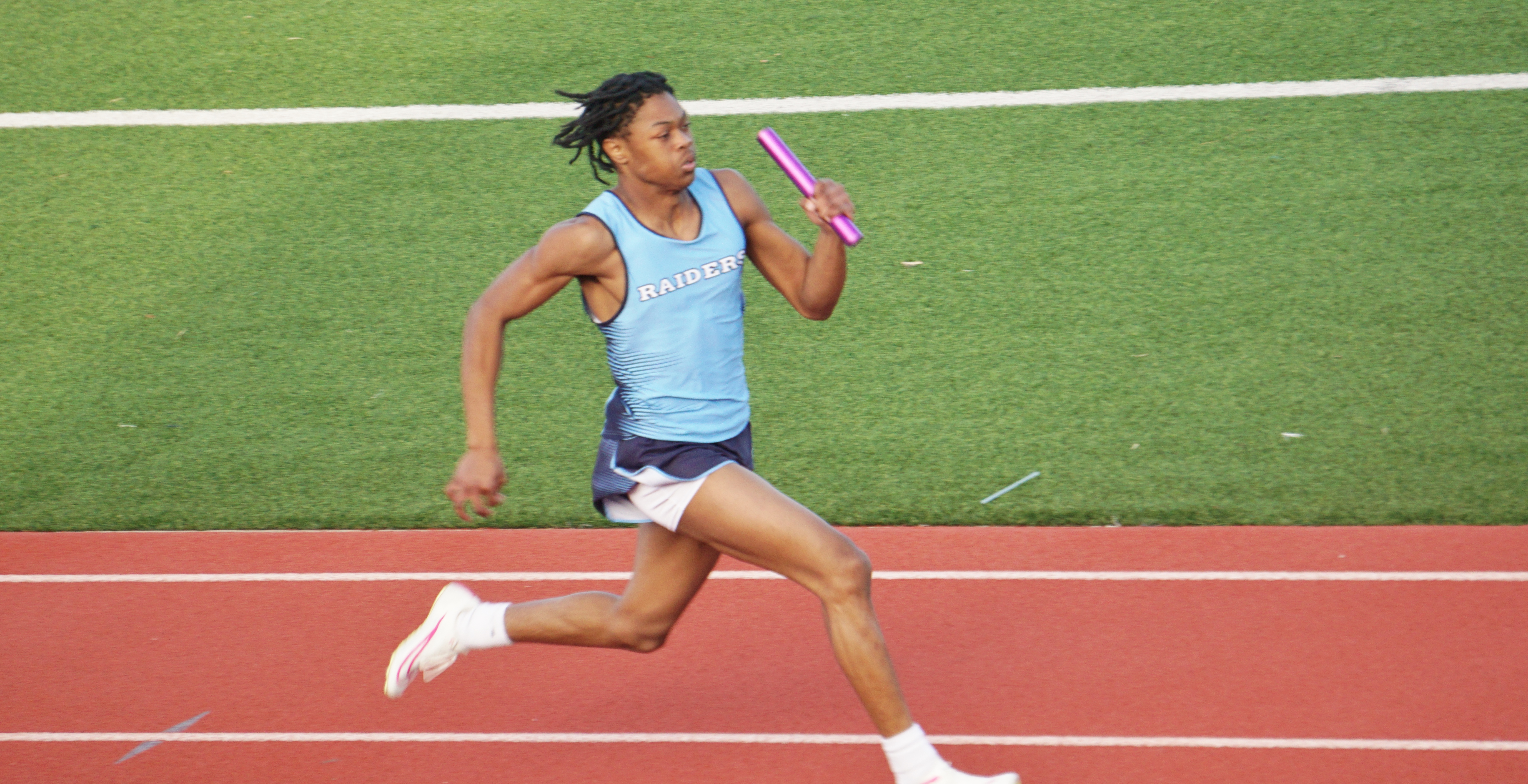 A female athlete in athletic attire is running on a red track surrounded by a green field.