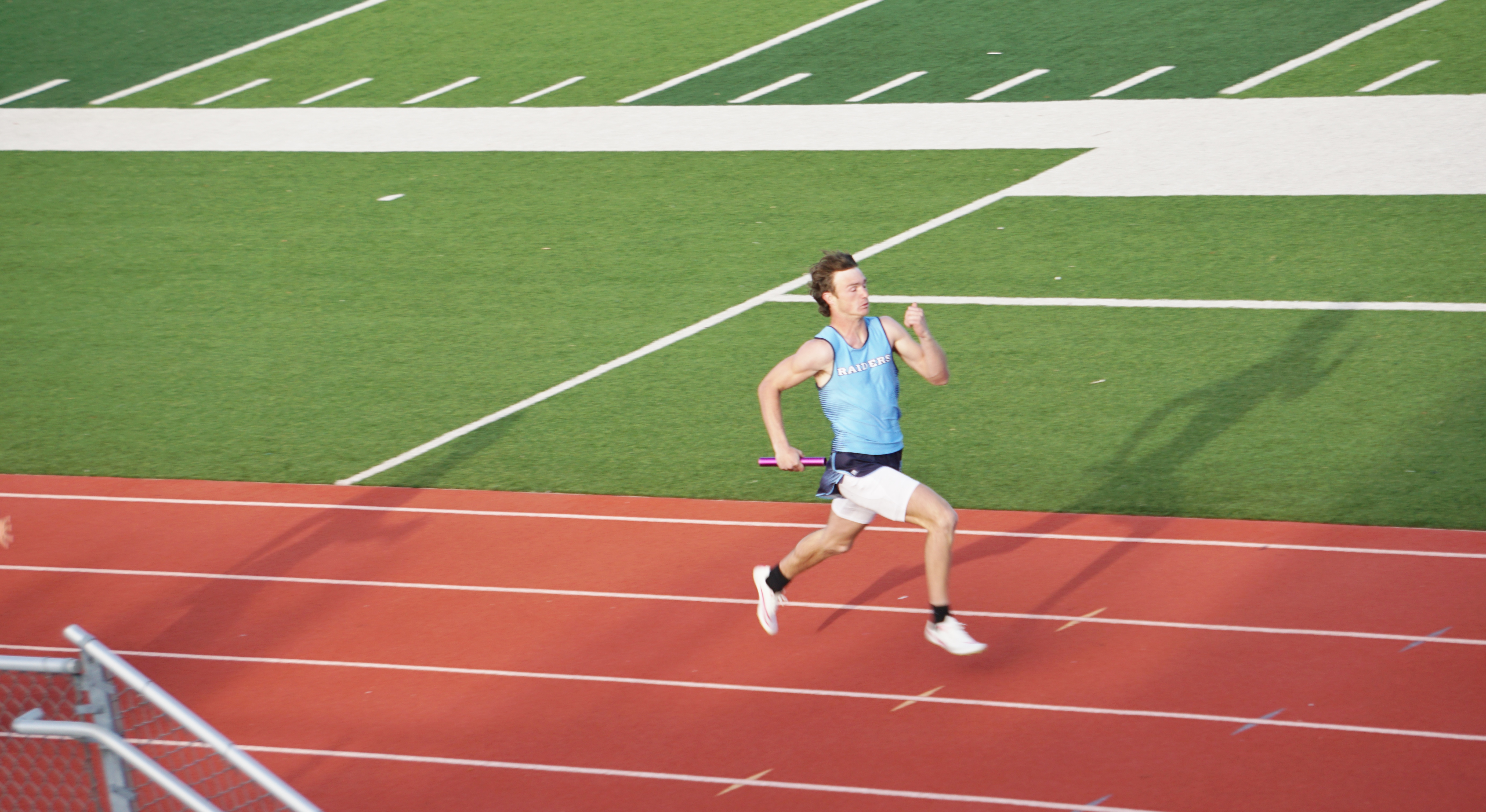 A person in a blue shirt and shorts is running on a red track surrounded by a green field and white lines.