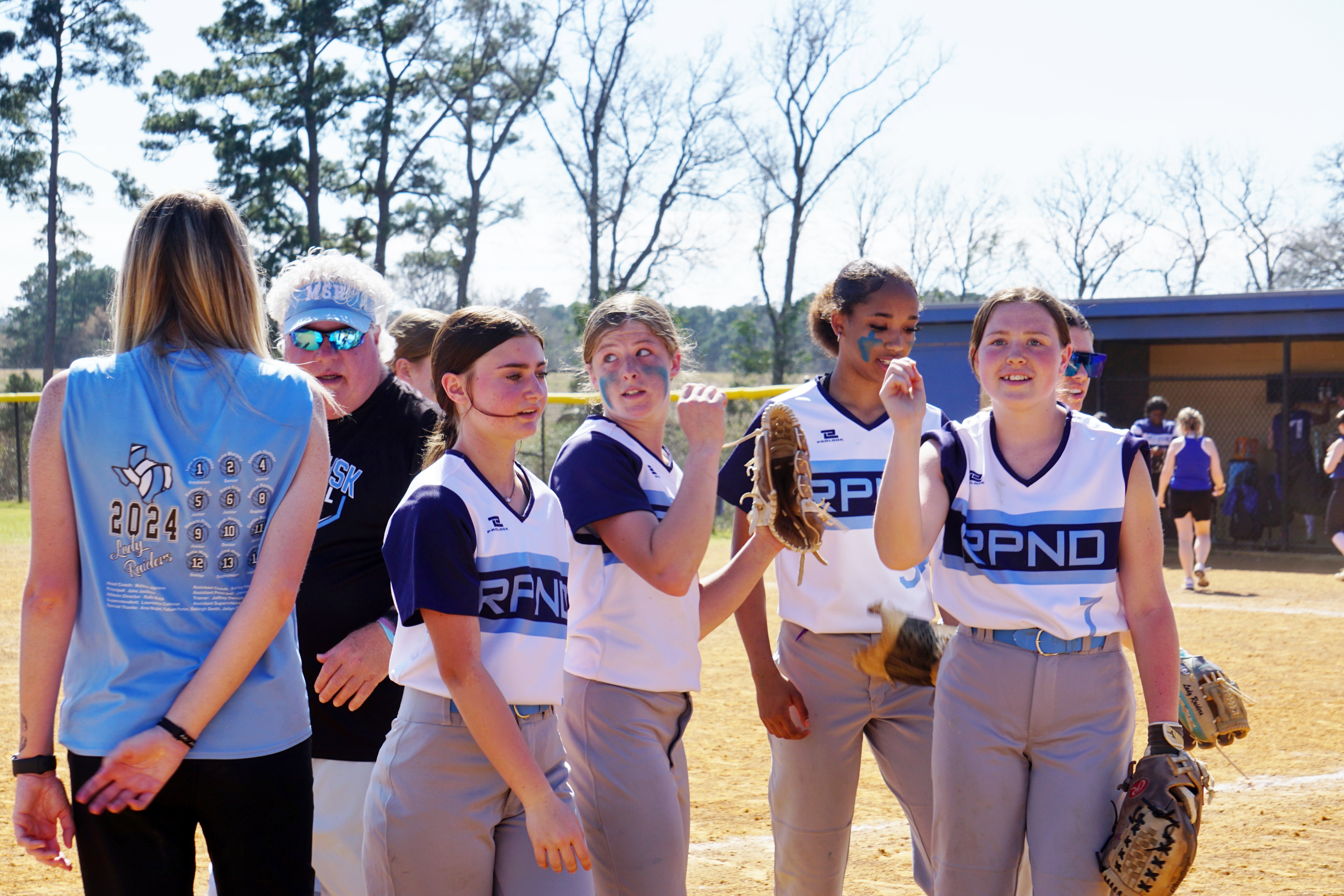 A group of young women in sports uniforms standing together on a baseball field surrounded by trees.