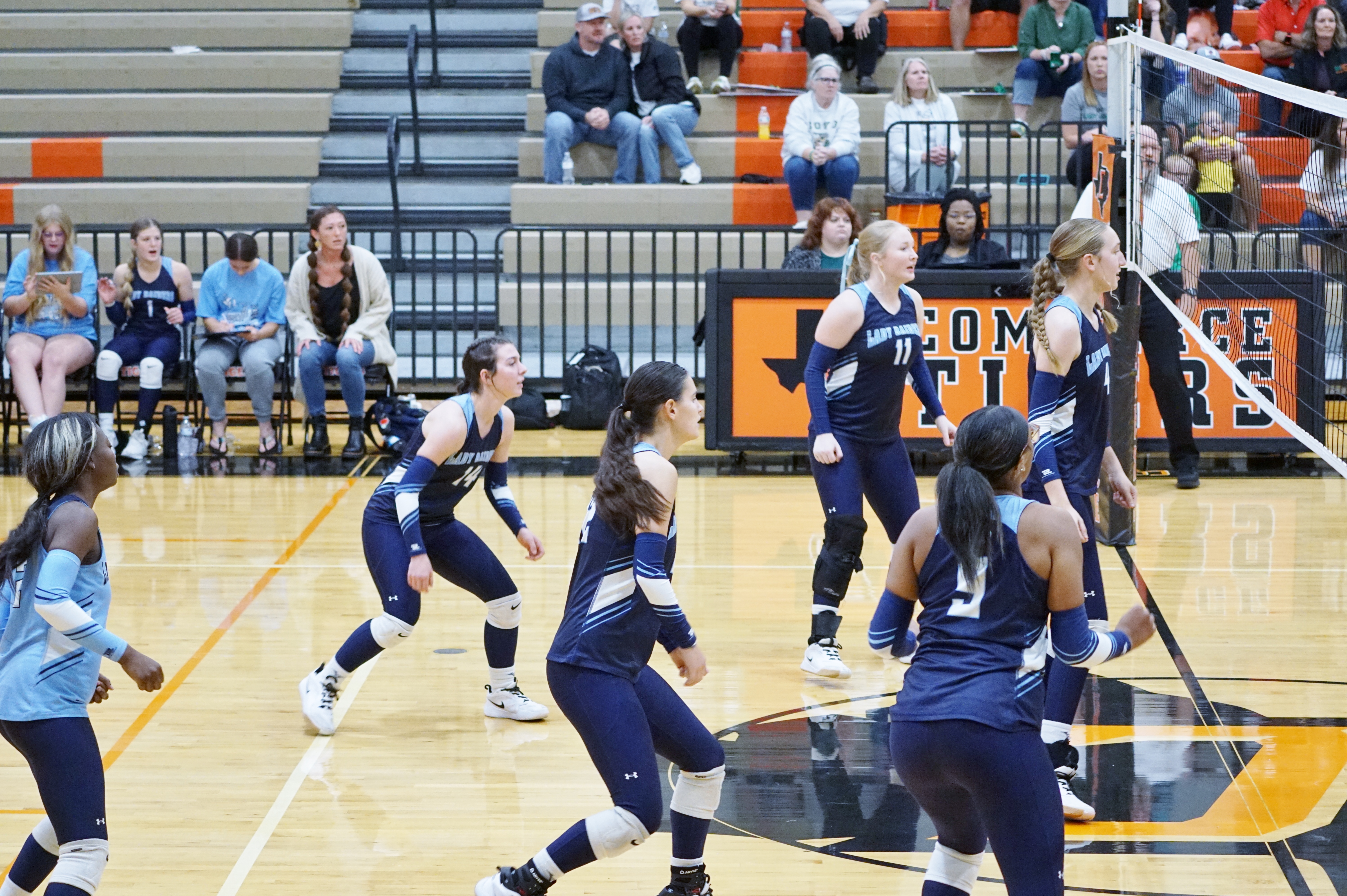 The image shows a group of people, likely a basketball team, on a basketball court during what appears to be a game or practice session. In the foreground, the players are wearing navy blue uniforms and are engaged in various basketball-related activities. In the background, there are spectators seated in the stands, suggesting this is taking place in a gymnasium or sports arena.