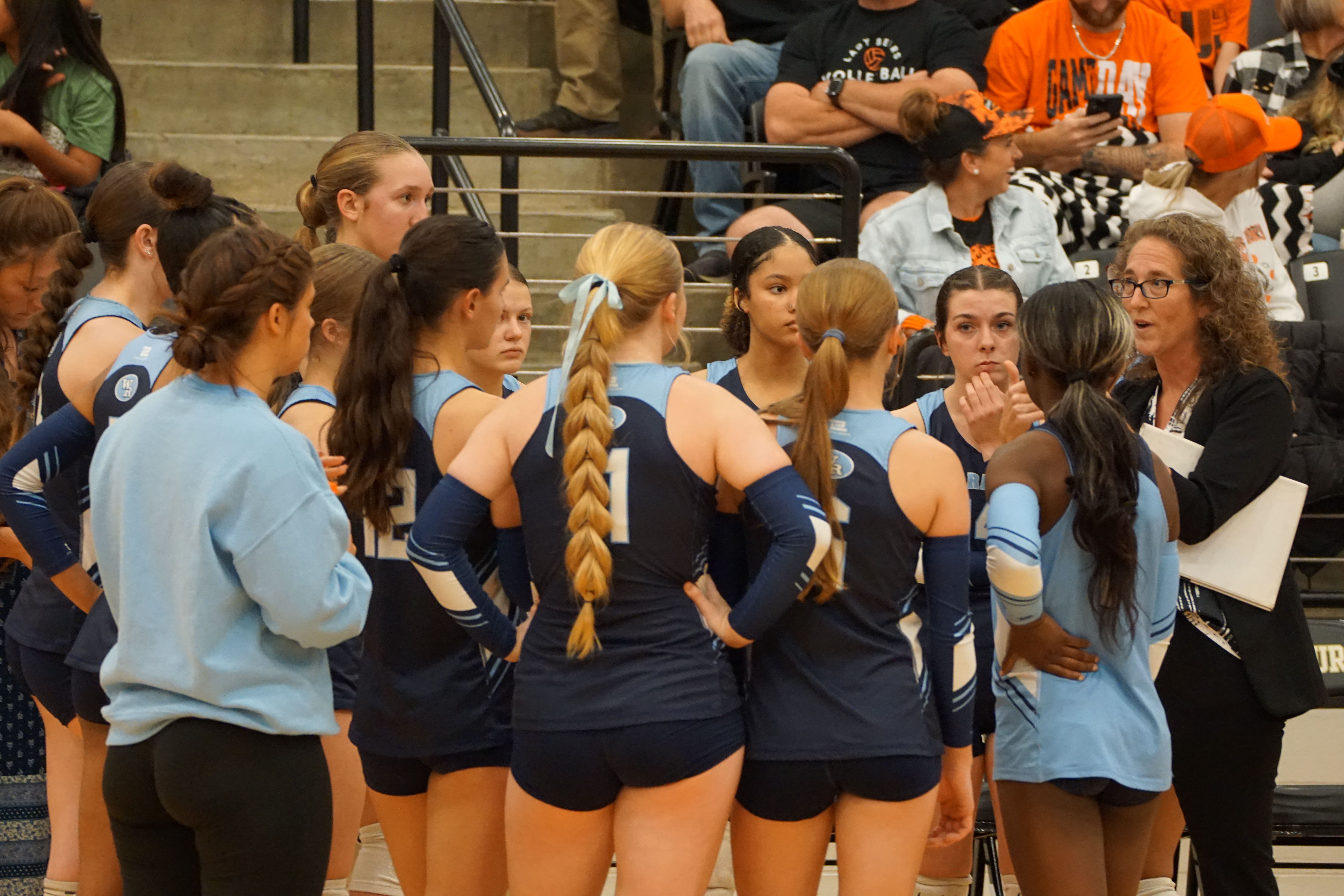 A group of young women in matching uniforms, likely cheerleaders or dancers, are gathered together in what appears to be a sports arena or event venue, with spectators visible in the background.
