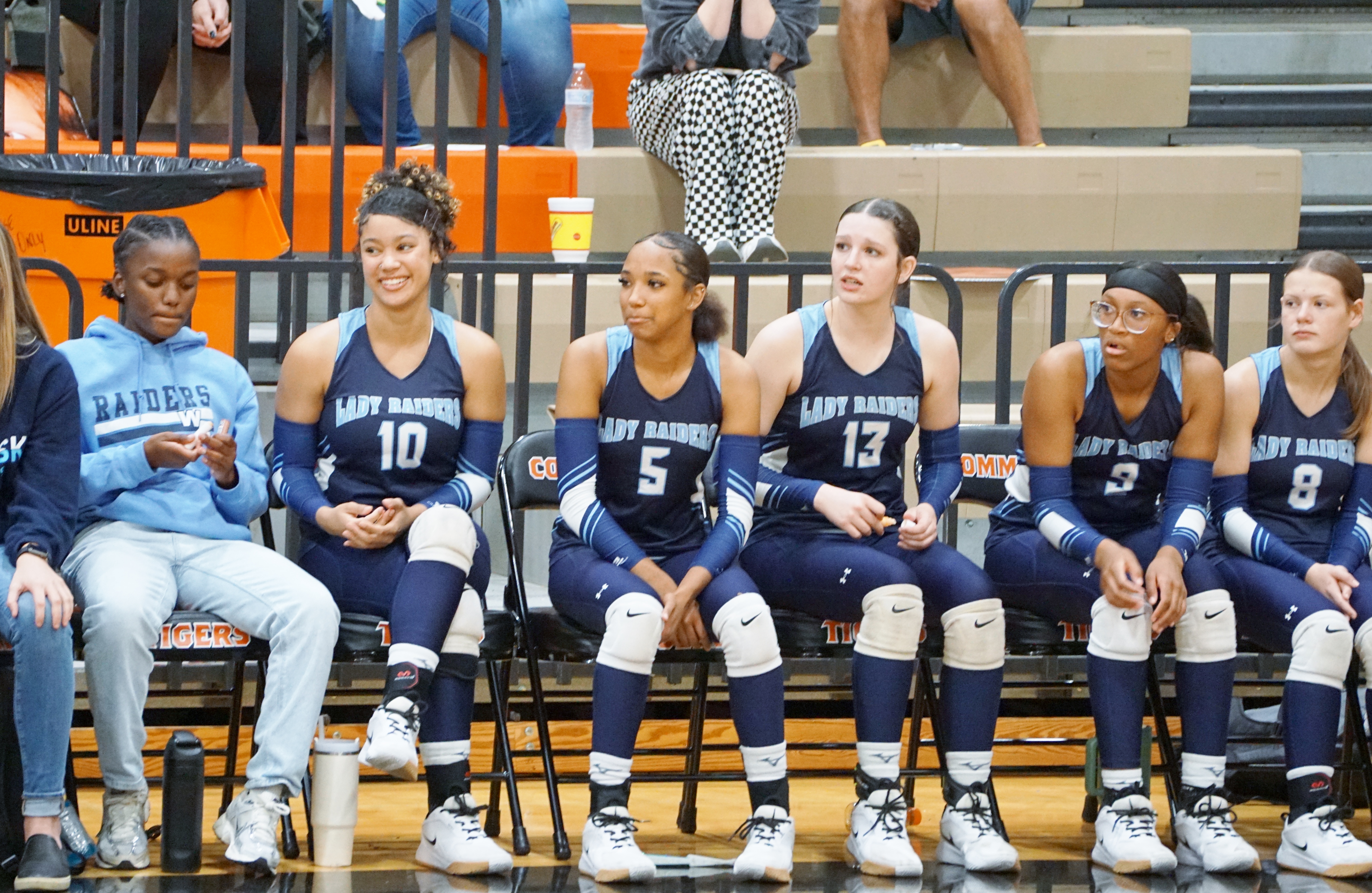 A group of female basketball players in navy blue uniforms sitting on a bench, with a spectator in the background and a basketball hoop visible.