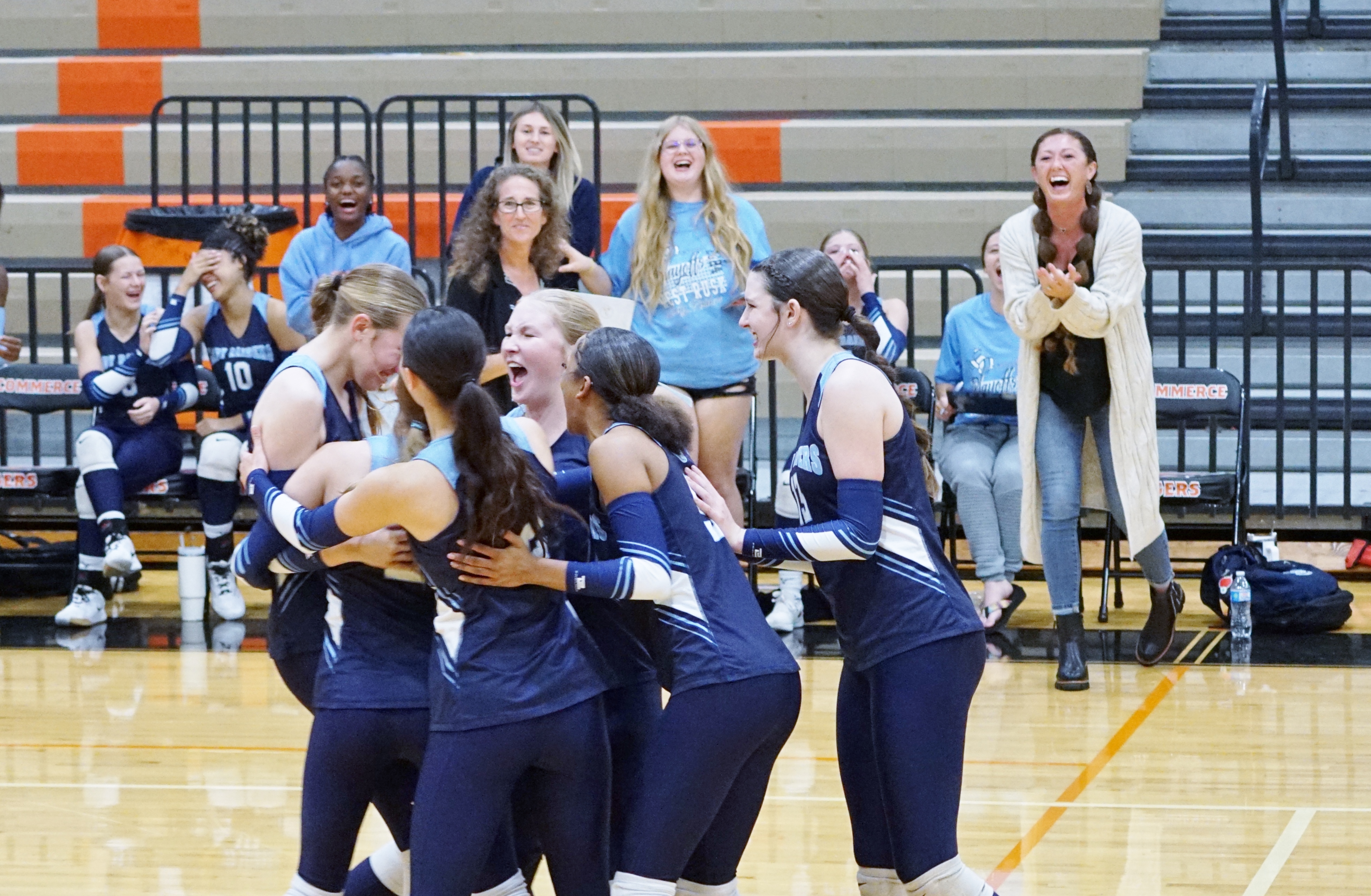 A group of cheerleaders in navy blue uniforms are gathered on a basketball court, surrounded by spectators in the stands behind them.