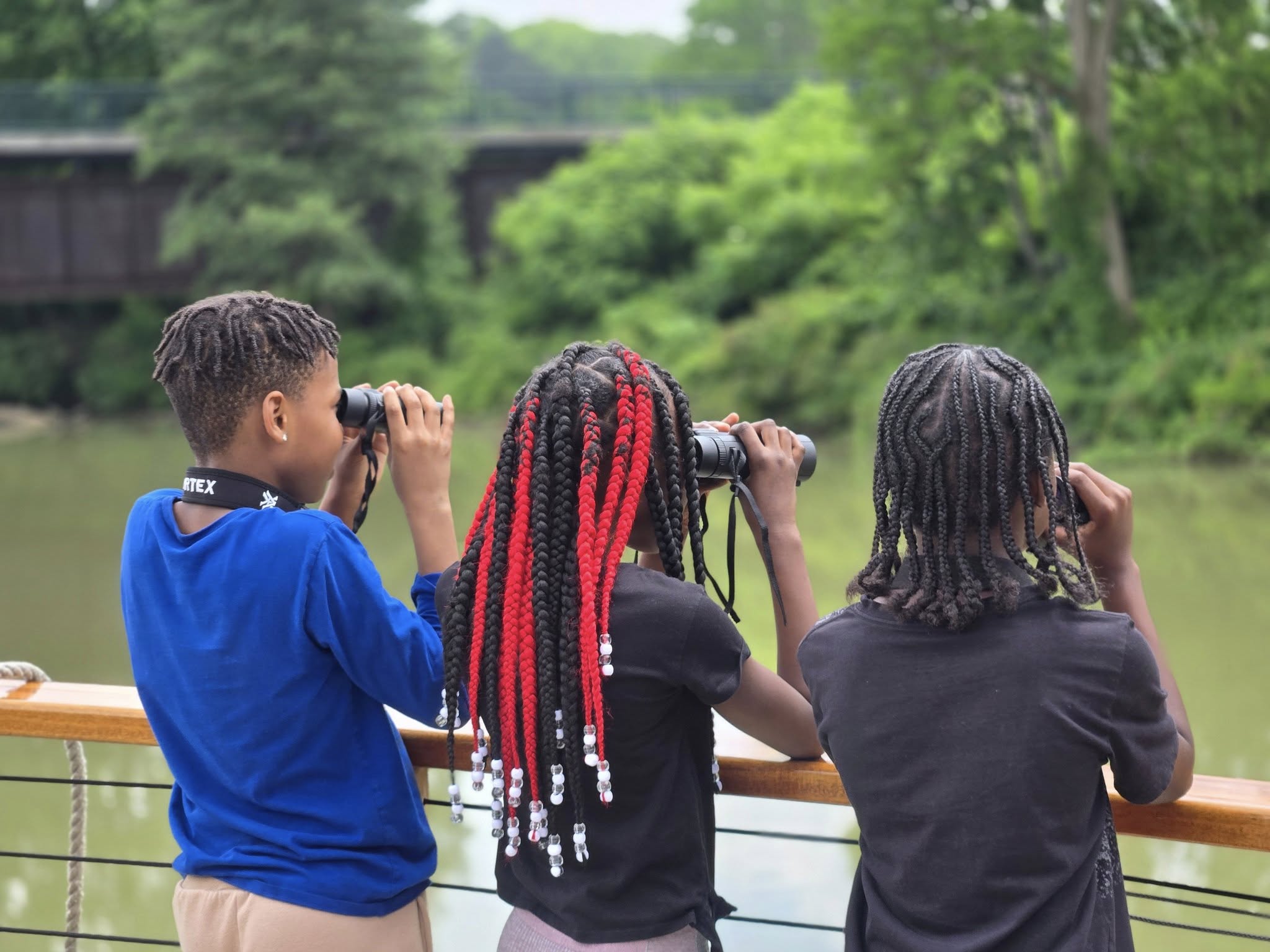 Three individuals with dreadlocked hairstyles are standing on a wooden deck, gazing through binoculars at the lush, green landscape in the background, which includes a body of water and a bridge.