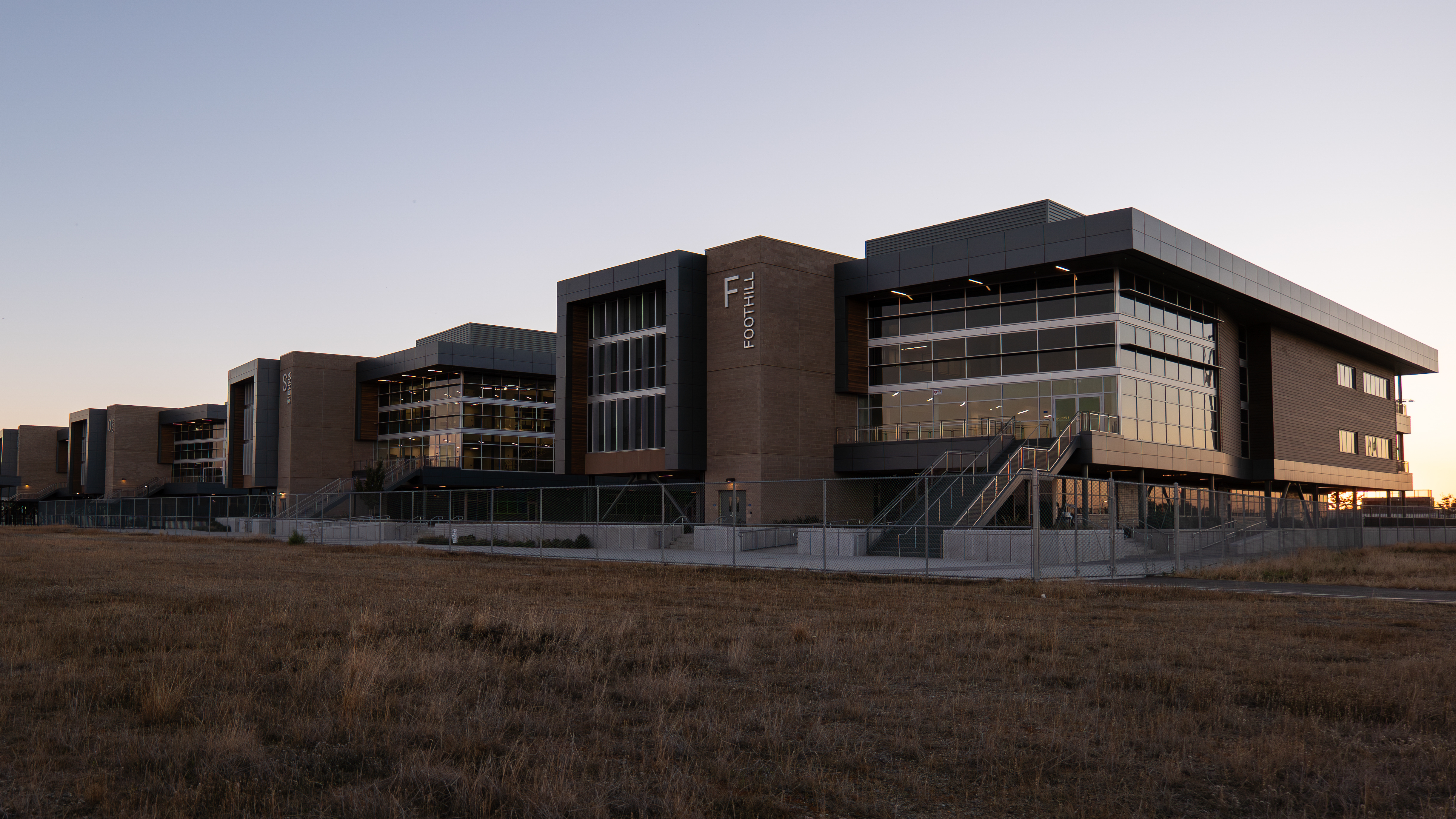 A modern, multi-story building complex stands in the foreground, surrounded by a grassy field in the background under a clear sky.