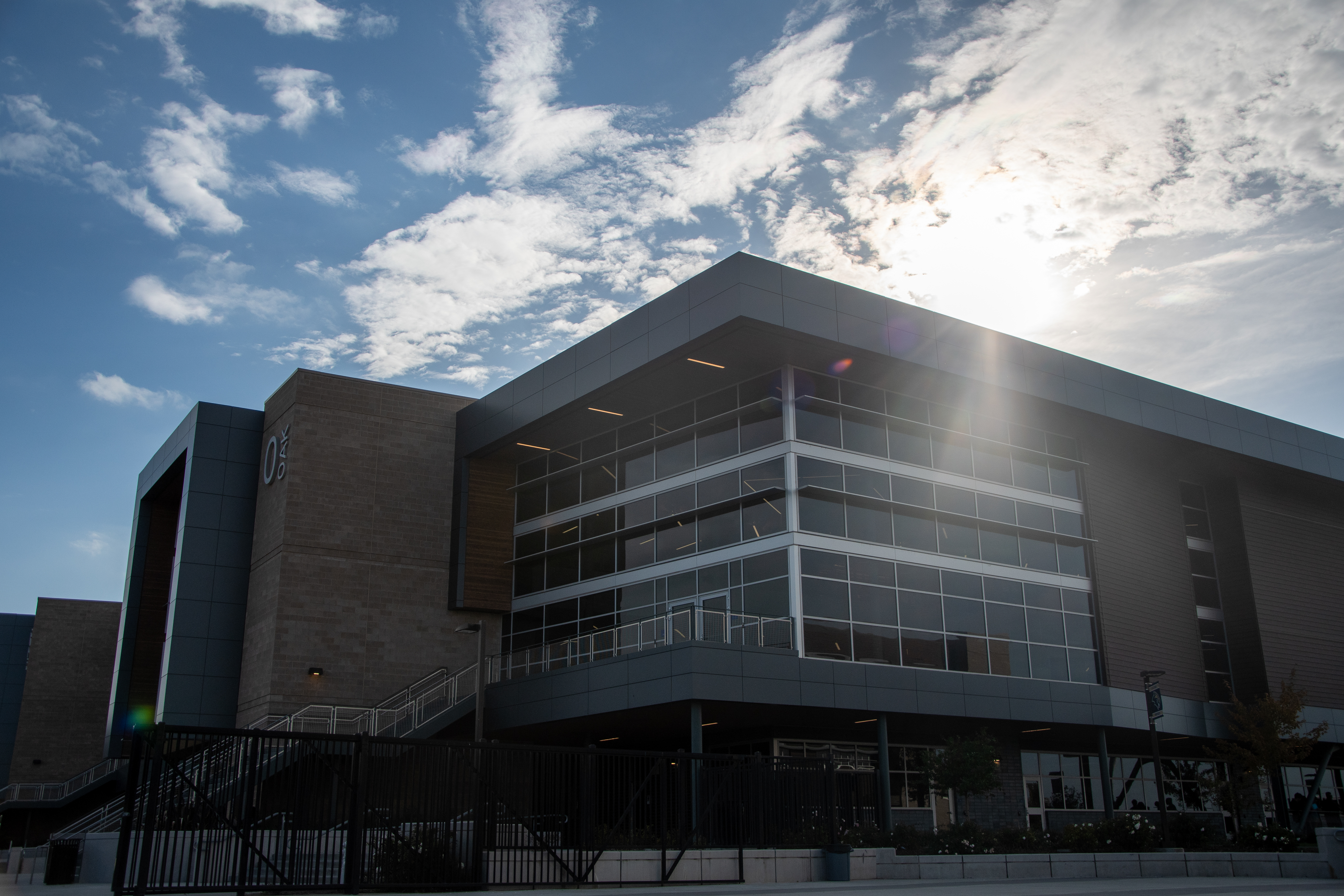A modern, multi-story building with a glass facade stands against a bright, cloudy sky, casting a shadow in the foreground.
