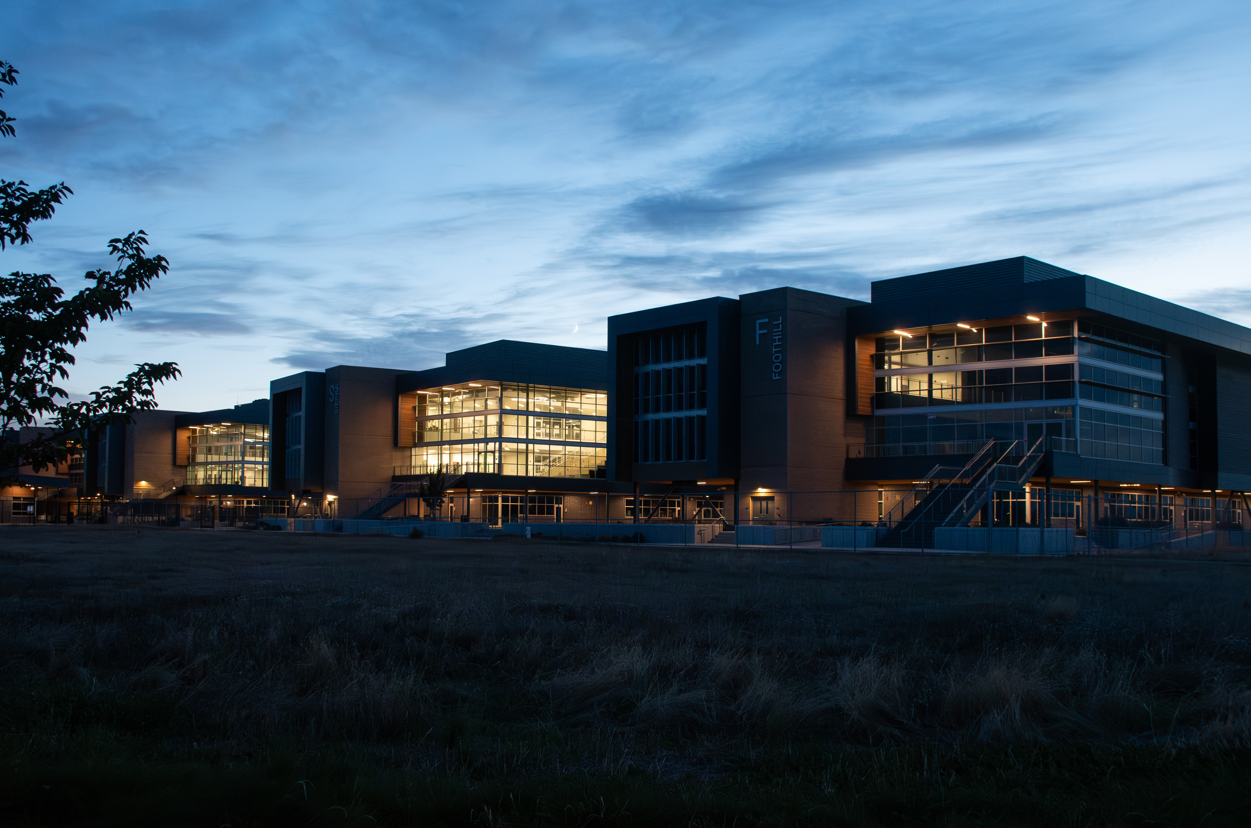 A modern, multi-story building with glass walls and illuminated interiors stands against a dark sky, surrounded by a grassy field in the foreground.
